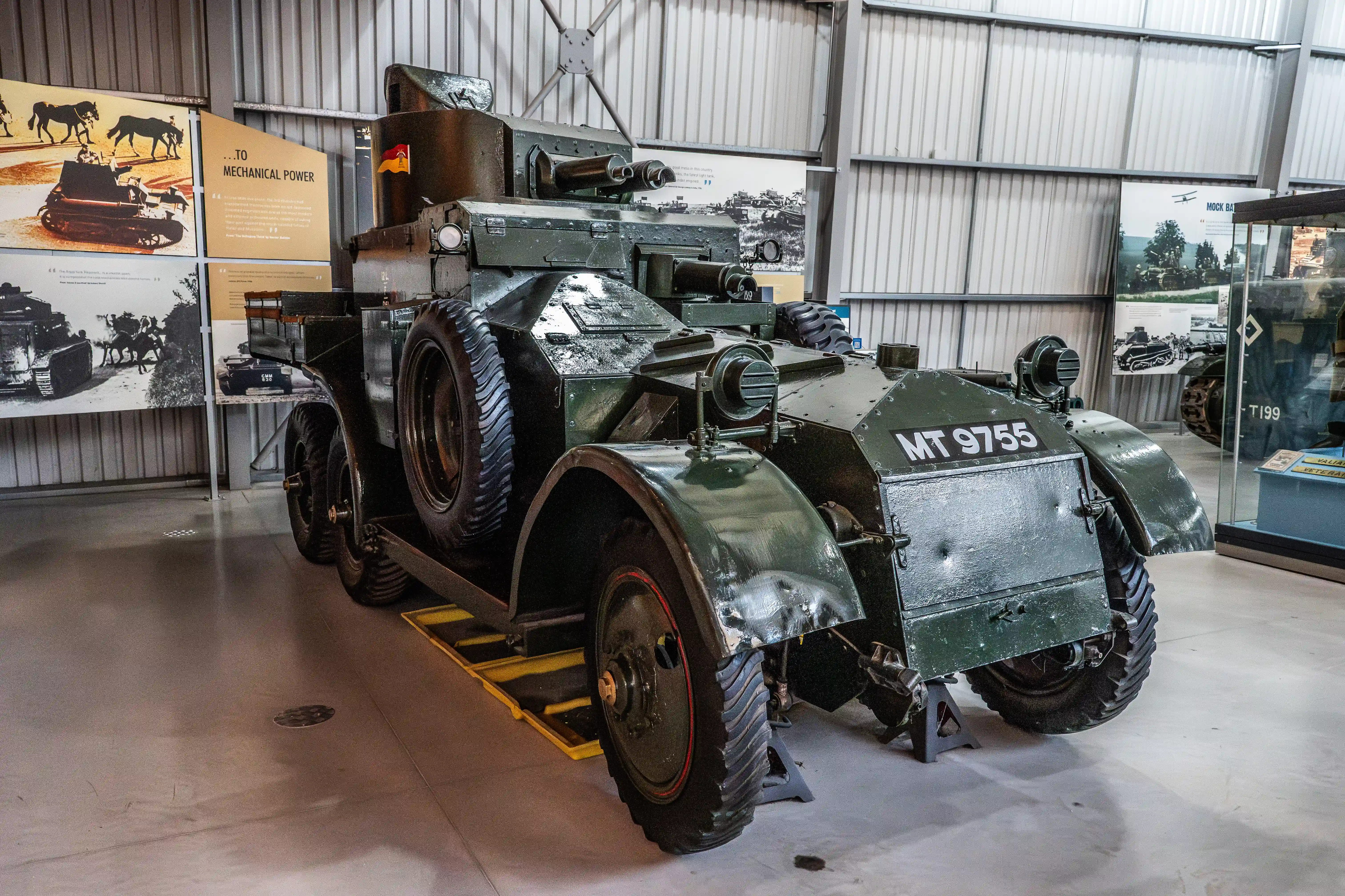 The image shows a vintage military vehicle, specifically an armored car, displayed in a museum setting. The vehicle has the designation 'MT 9755' marked on its side and features large, rugged tires and a heavily armored body. The display area includes informational panels with historical photographs and text, providing context about the transition from horse-drawn vehicles to mechanized ones. The setting appears to be well-lit and organized, with the vehicle positioned on a raised platform for viewing.
