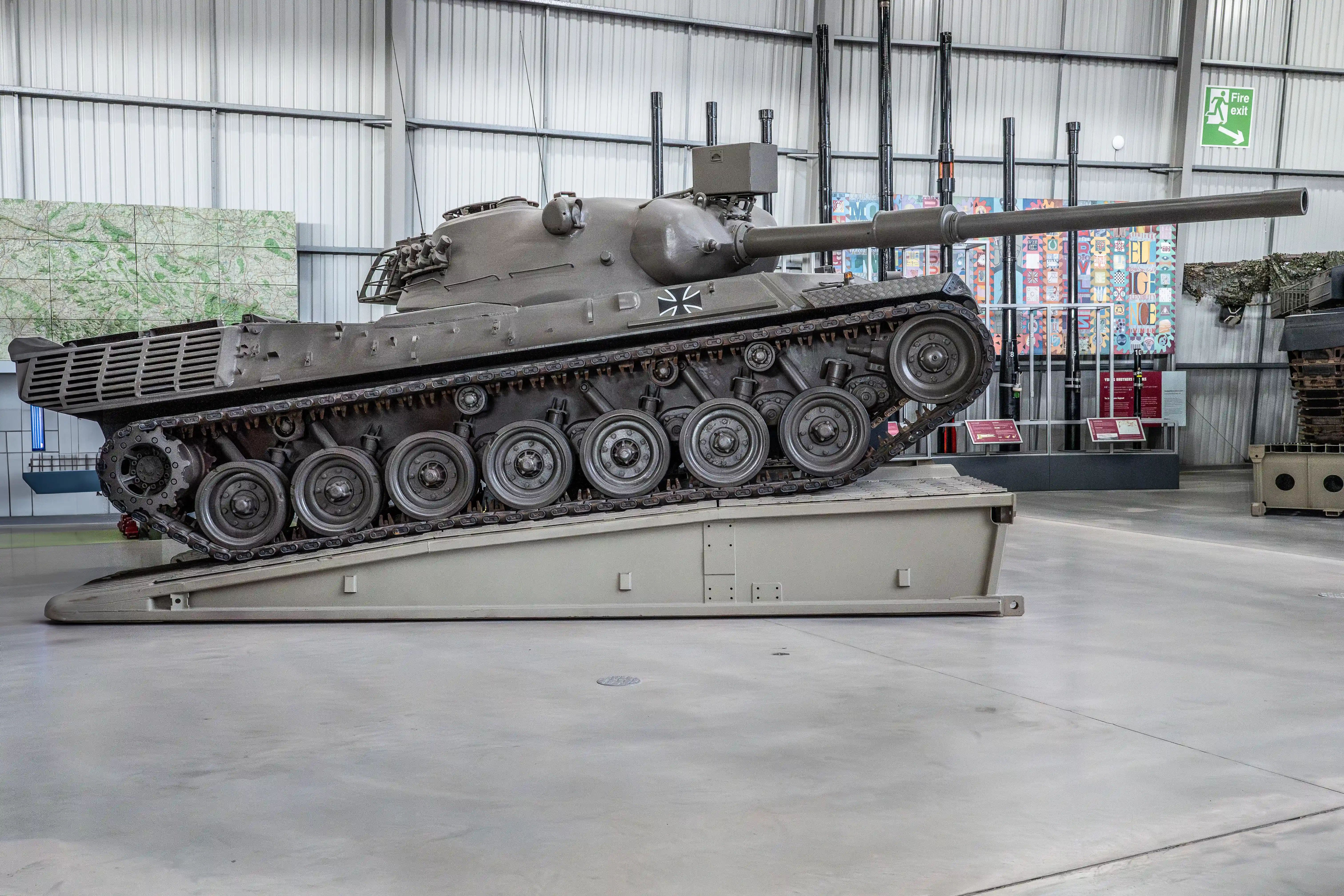 The image depicts a tank displayed in a museum setting. The tank is elevated on a platform, showcasing its tracks and underbelly. The environment appears to be indoors with a large, open space and high ceilings. There are informational placards near the tank, likely providing details about its history and specifications. The overall setting suggests an educational exhibit focused on military history.