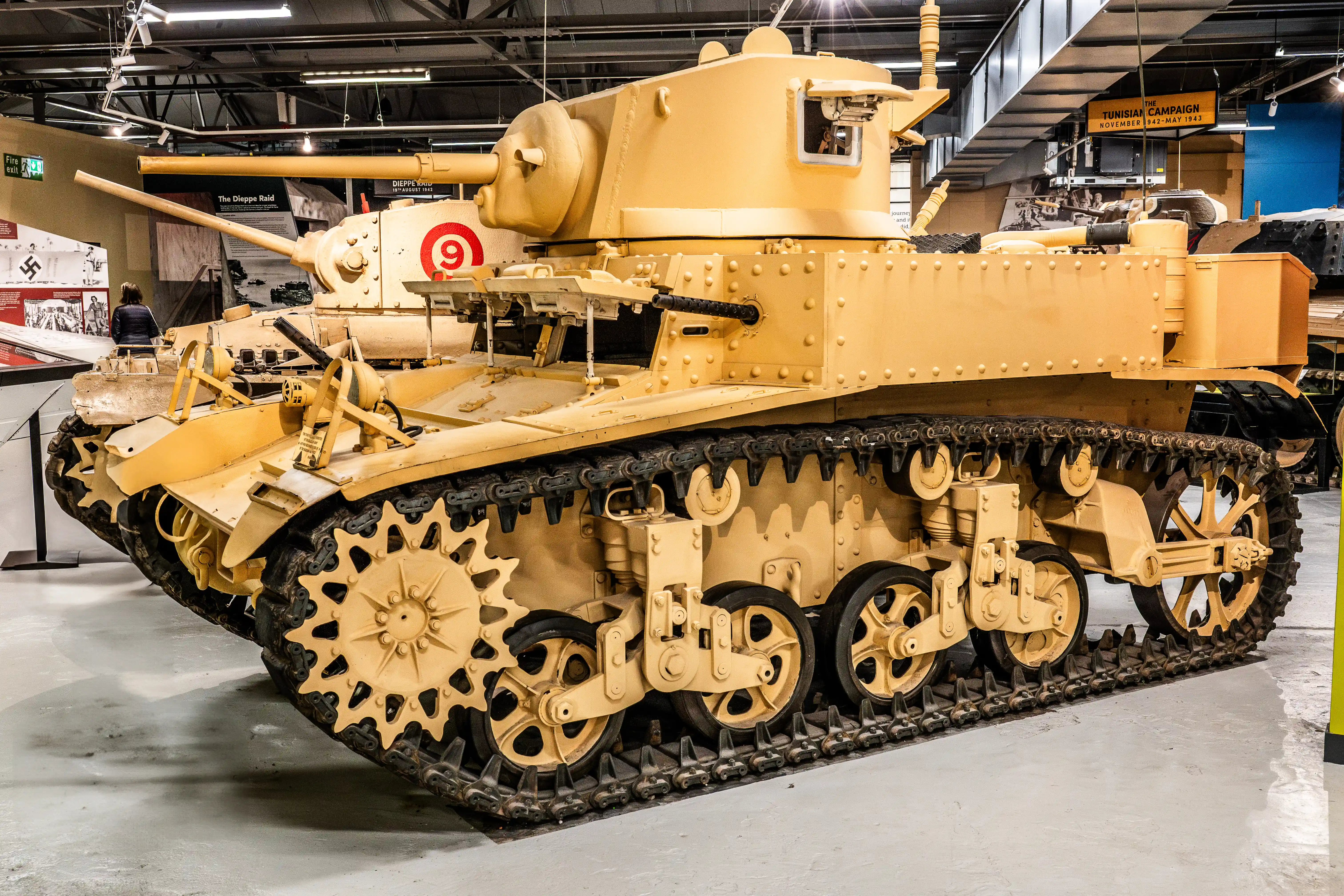 The image depicts a historical military tank on display in a museum. The tank is painted in a sandy yellow color and has the number 9 marked on its side. It features a large turret with a prominent gun barrel, and its tracks and wheels are visible, showcasing its robust and rugged design. The background includes various informational displays and banners, indicating that the tank is part of an exhibit related to historical military campaigns, specifically mentioning the Tunisian Campaign from November 1942 to May 1943.