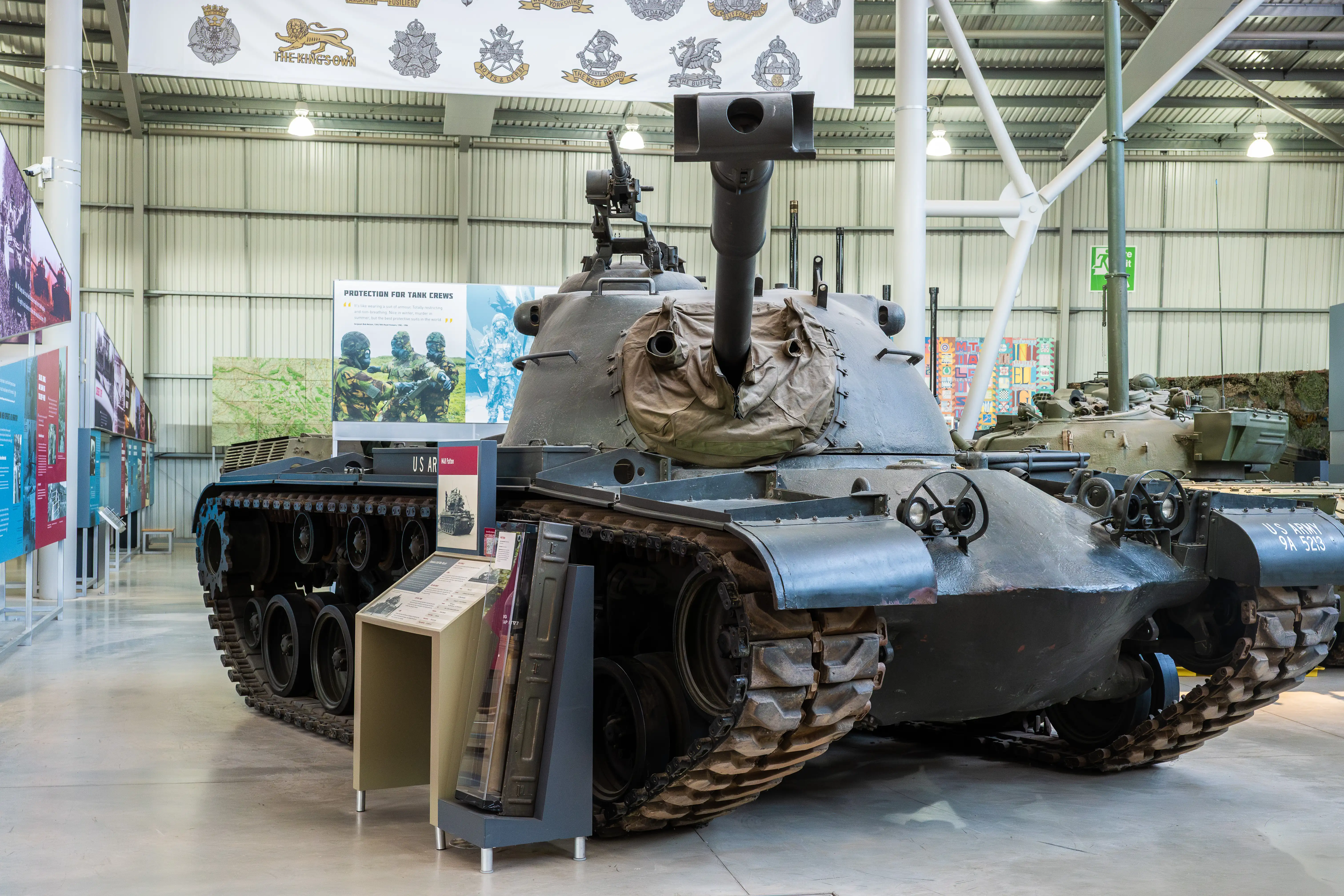 The image depicts a military tank displayed in a museum. The tank is surrounded by informational placards and is housed in a large, well-lit exhibition hall. Various military insignia and banners are visible on the ceiling, and other military vehicles and exhibits can be seen in the background.