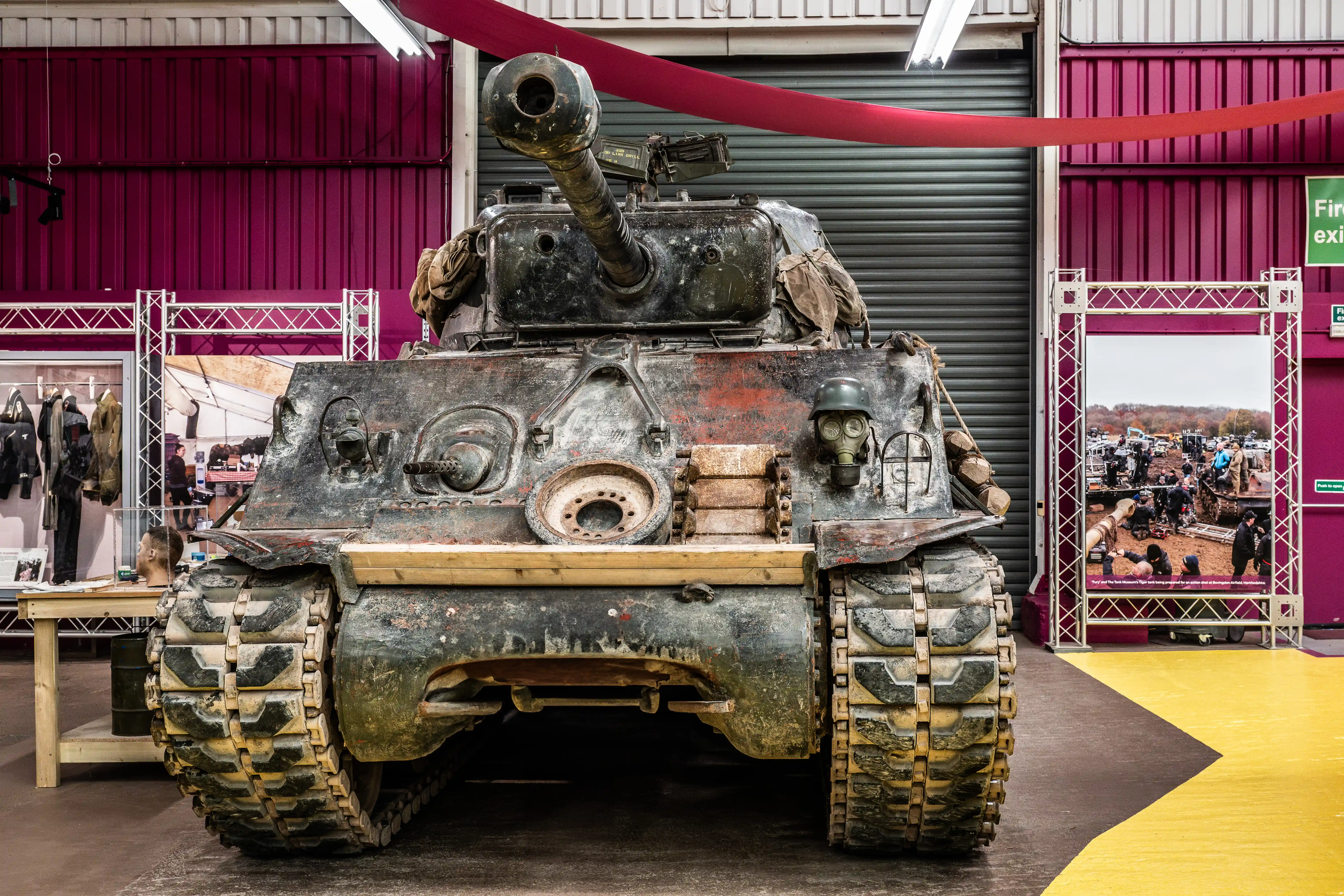 The image depicts a vintage military tank on display at an indoor exhibition. The tank is weathered and adorned with camouflage netting and various equipment, including a mannequin wearing a gas mask. The exhibition space features red and yellow flooring, with other displays and attendees visible in the background.