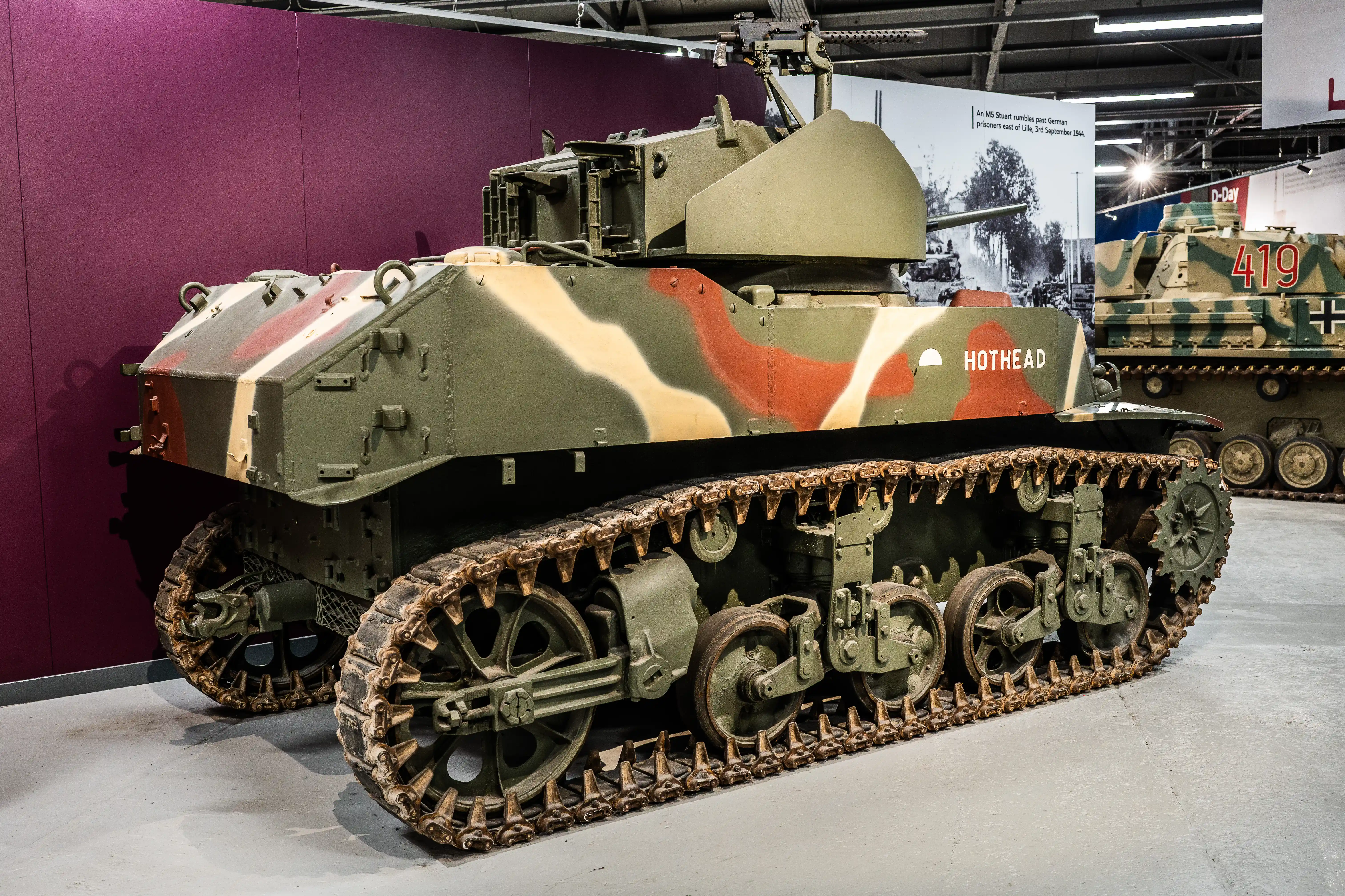 The image shows a military tank on display, likely in a museum setting. The tank is painted in a camouflage pattern of green, brown, and black. The name 'HOTHEAD' is prominently displayed on the side of the tank. The tank has a turret with a mounted machine gun and is equipped with continuous tracks for mobility. In the background, another tank is visible, marked with the number '419'.