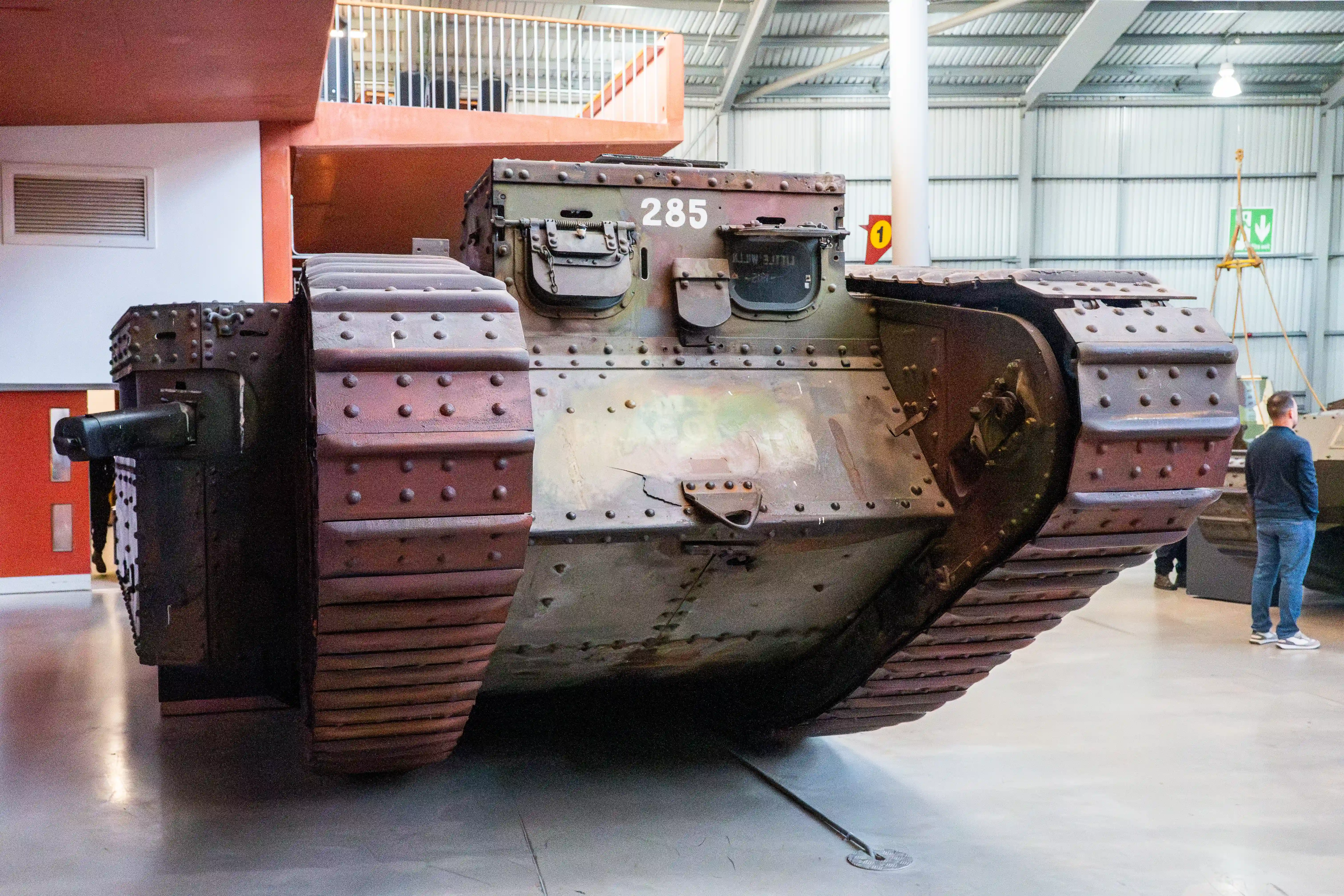 The image shows a vintage military tank on display in a museum. The tank, marked with the number 285, features riveted armor plating and a turret with a mounted gun. The setting appears to be an indoor exhibit area with a high ceiling and industrial-style architecture. A person is seen observing the tank, providing a sense of scale to the size of the vehicle.
