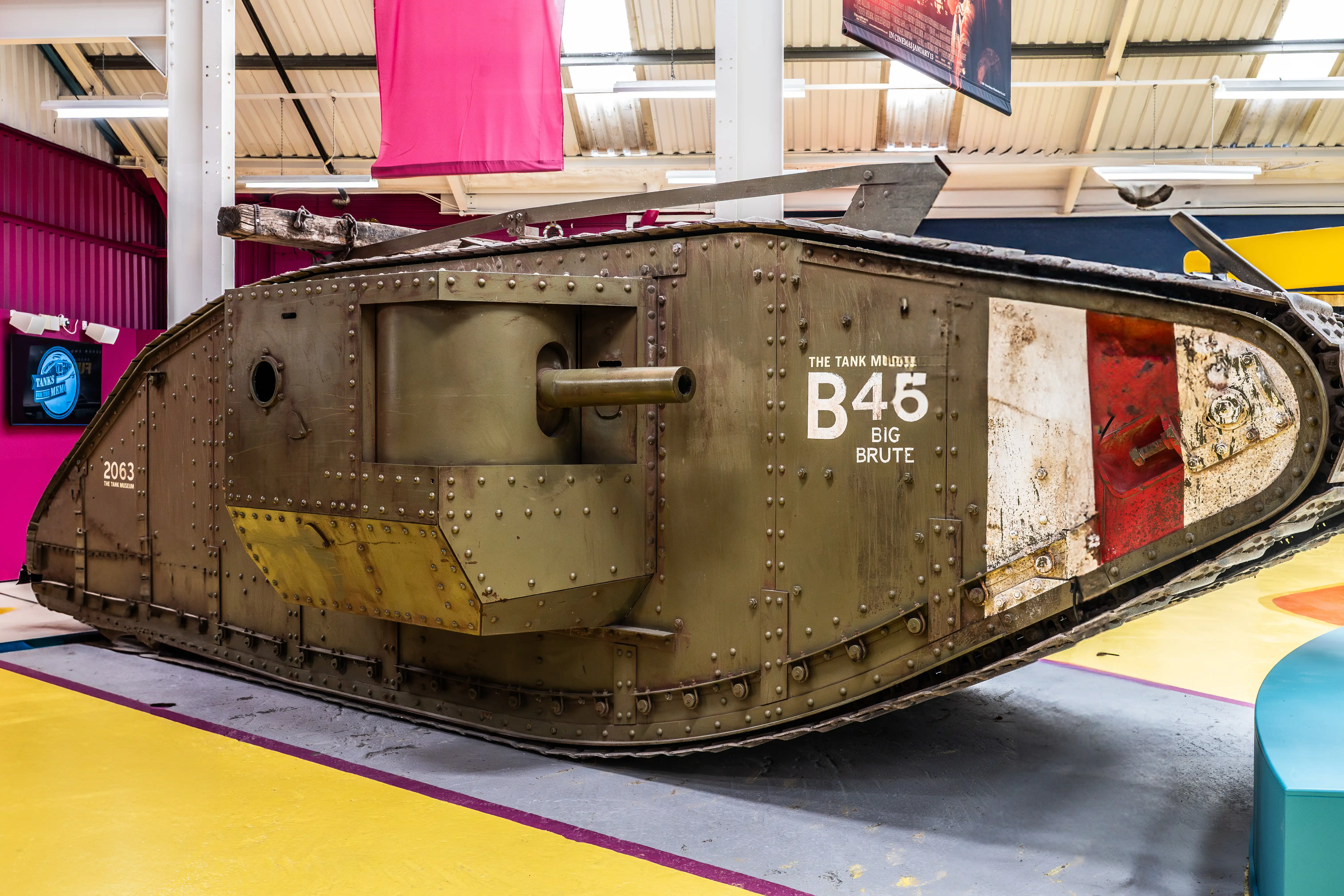 The image shows a historical military tank, specifically a British Mark IV tank used during World War I. The tank is displayed indoors, likely in a museum setting. It has a rugged, boxy structure with riveted armor plating and a prominent central barrel. The tank is labeled with 'B45 Big Brute' and 'The Tank Museum' along with the number '2063'. The surrounding area is colorful, suggesting an educational or interactive exhibit.