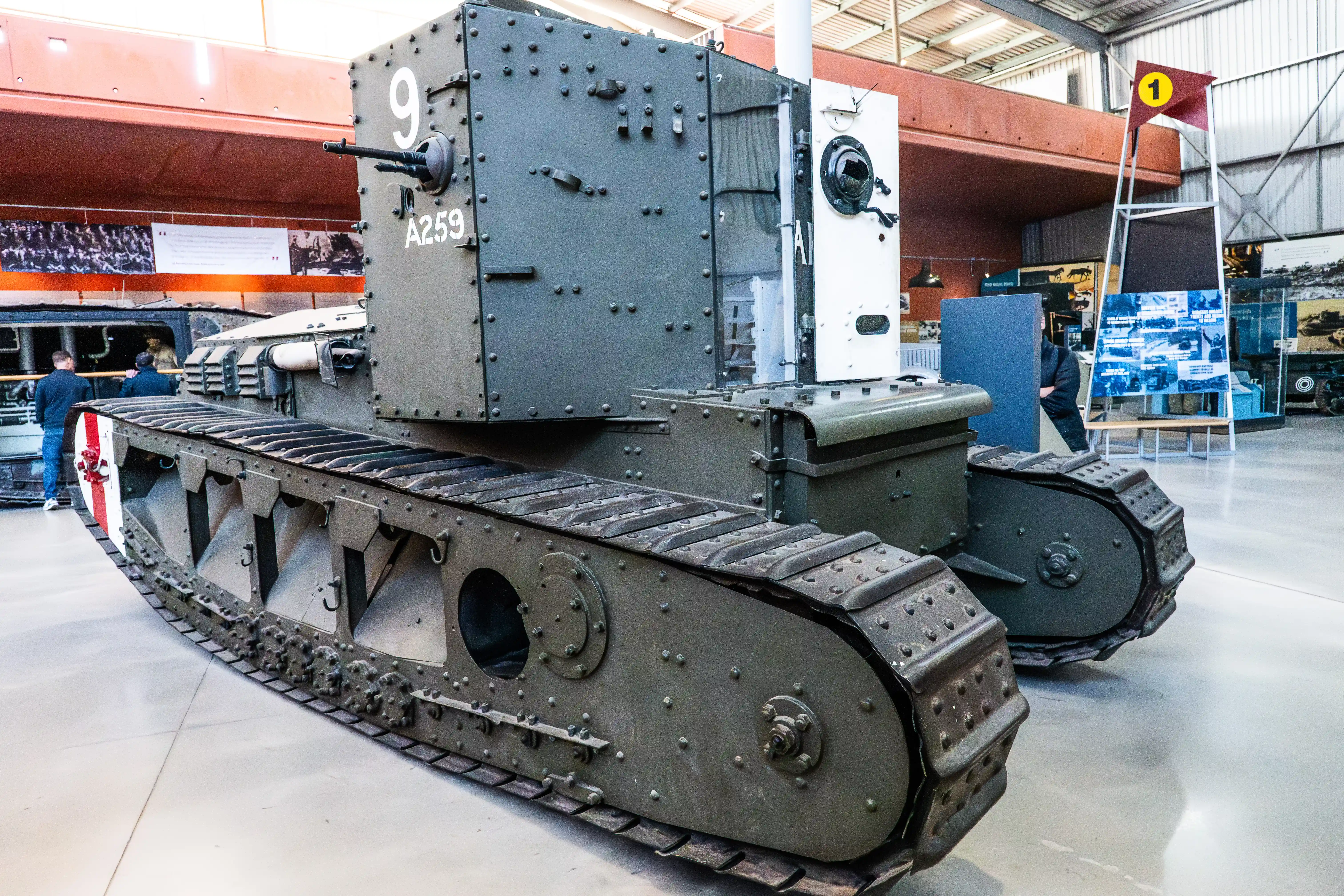 The image depicts a vintage tank on display in a museum. The tank, marked with the number 9 and the identifier A259, features riveted armor and continuous track links characteristic of early 20th-century tank designs. The museum setting includes various informational displays and other military artifacts, providing educational context about the historical significance and technical details of the tank.