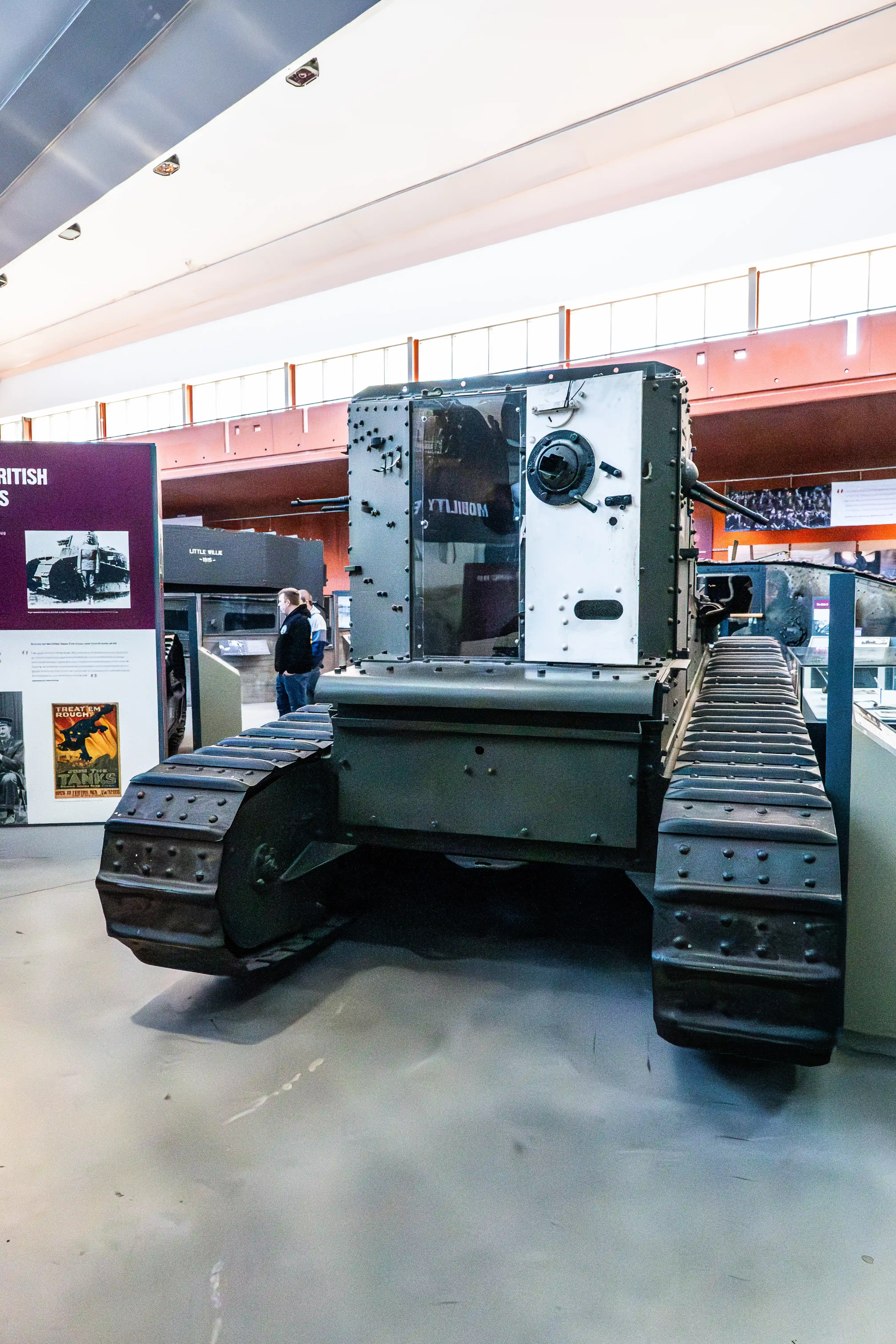 The image shows a historical tank on display in a museum. The tank has a robust, armored structure with tracks designed for rugged terrain. It is positioned in an indoor exhibit area with informational displays in the background, suggesting it is part of a larger collection or exhibition related to military history. The setting appears to be well-lit and spacious, with other exhibits and visitors visible in the background.