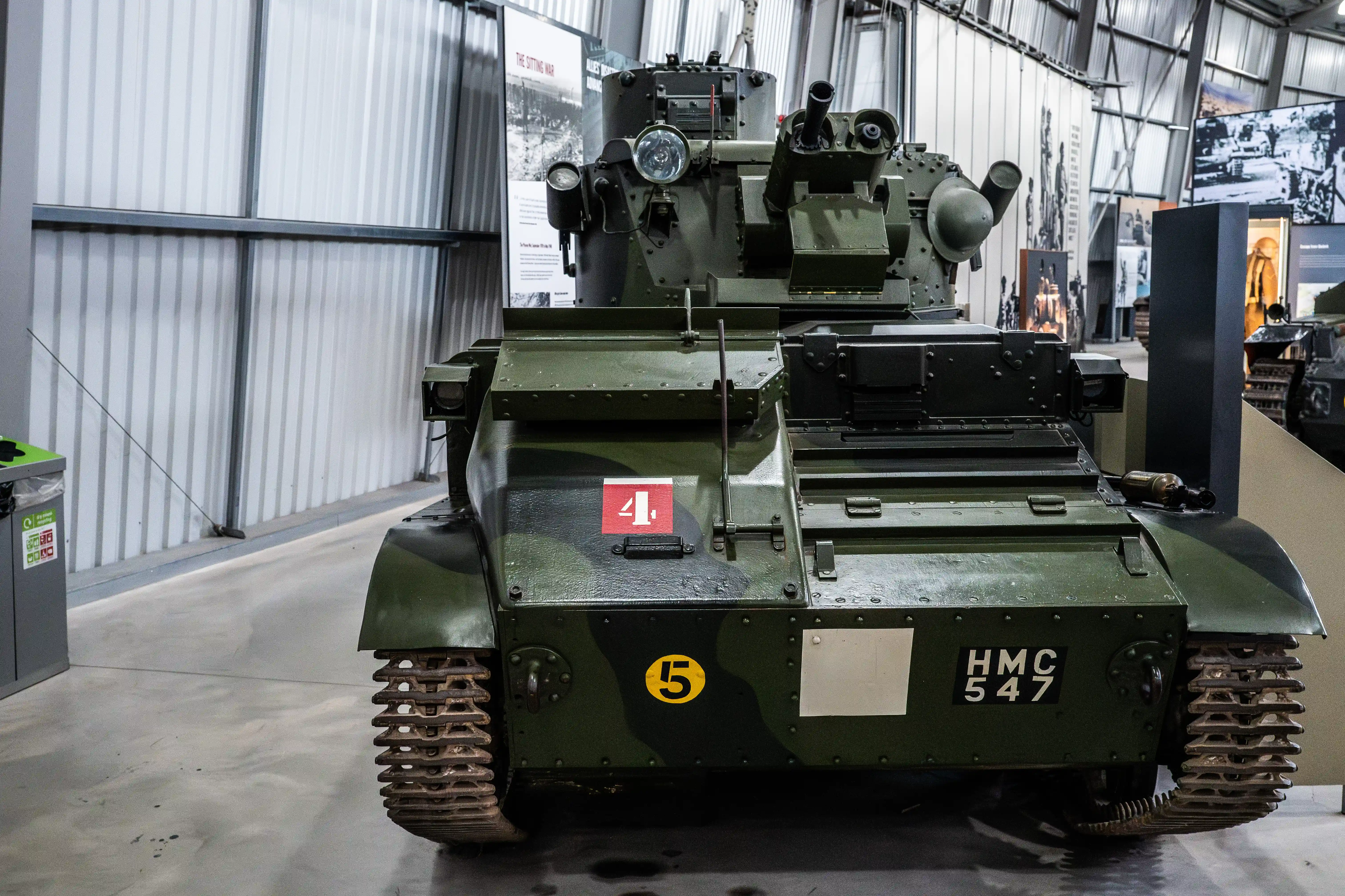The image depicts a military tank on display in a museum or exhibition setting. The tank is painted in a green camouflage pattern and has various markings, including the number 41 on a red background and HMC 547 on the front. It is equipped with a turret-mounted machine gun and other military equipment. The setting appears to be indoors, with informational displays and other military artifacts visible in the background.