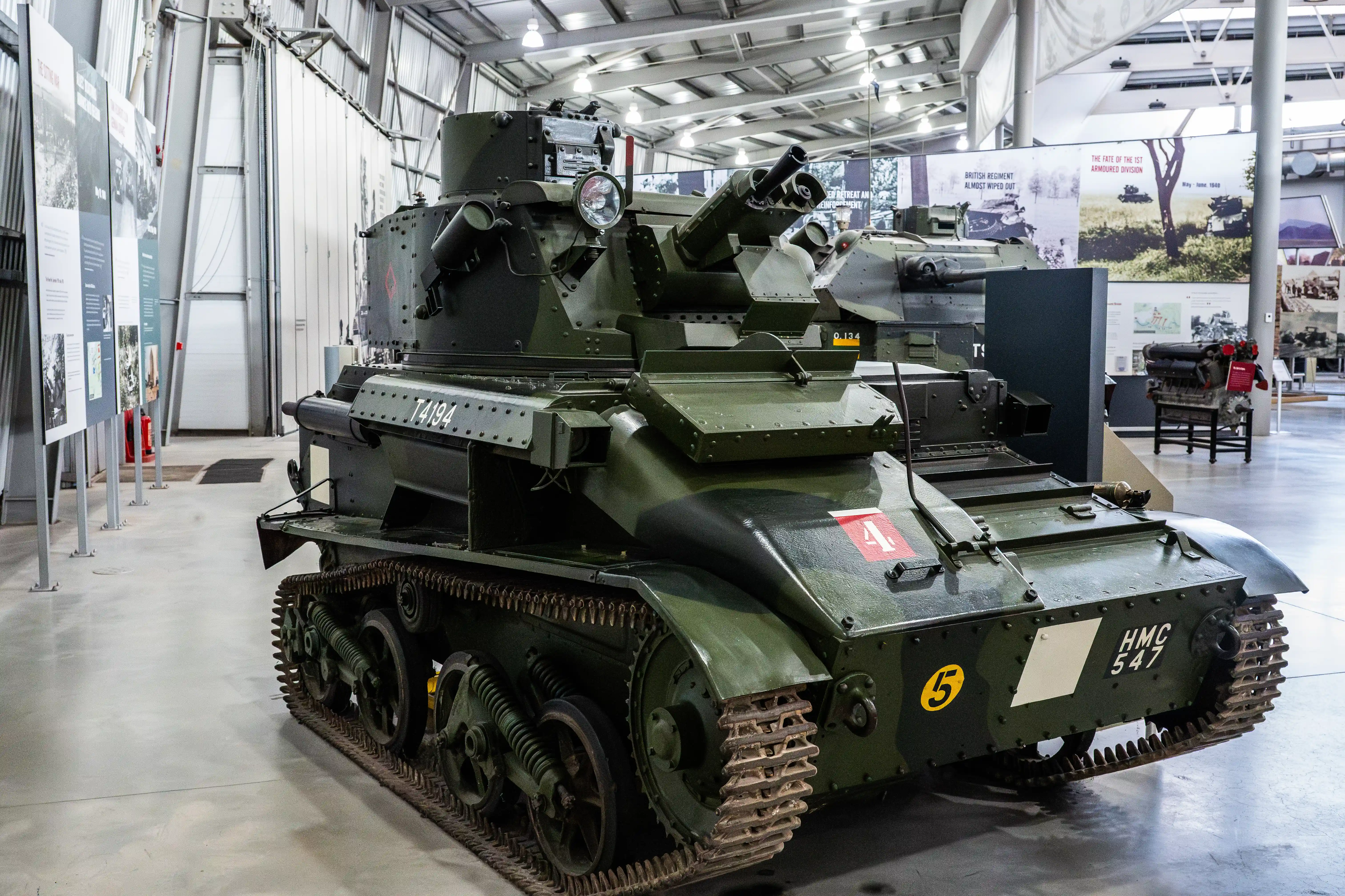 The image depicts a vintage military tank on display in a museum. The tank is green and has caterpillar tracks, with various markings and numbers on its body, including 'T4194' and 'HMC 547'. It is equipped with a turret mounted on top, which houses a large gun. The museum setting is evident from the informational displays and other military artifacts visible in the background.