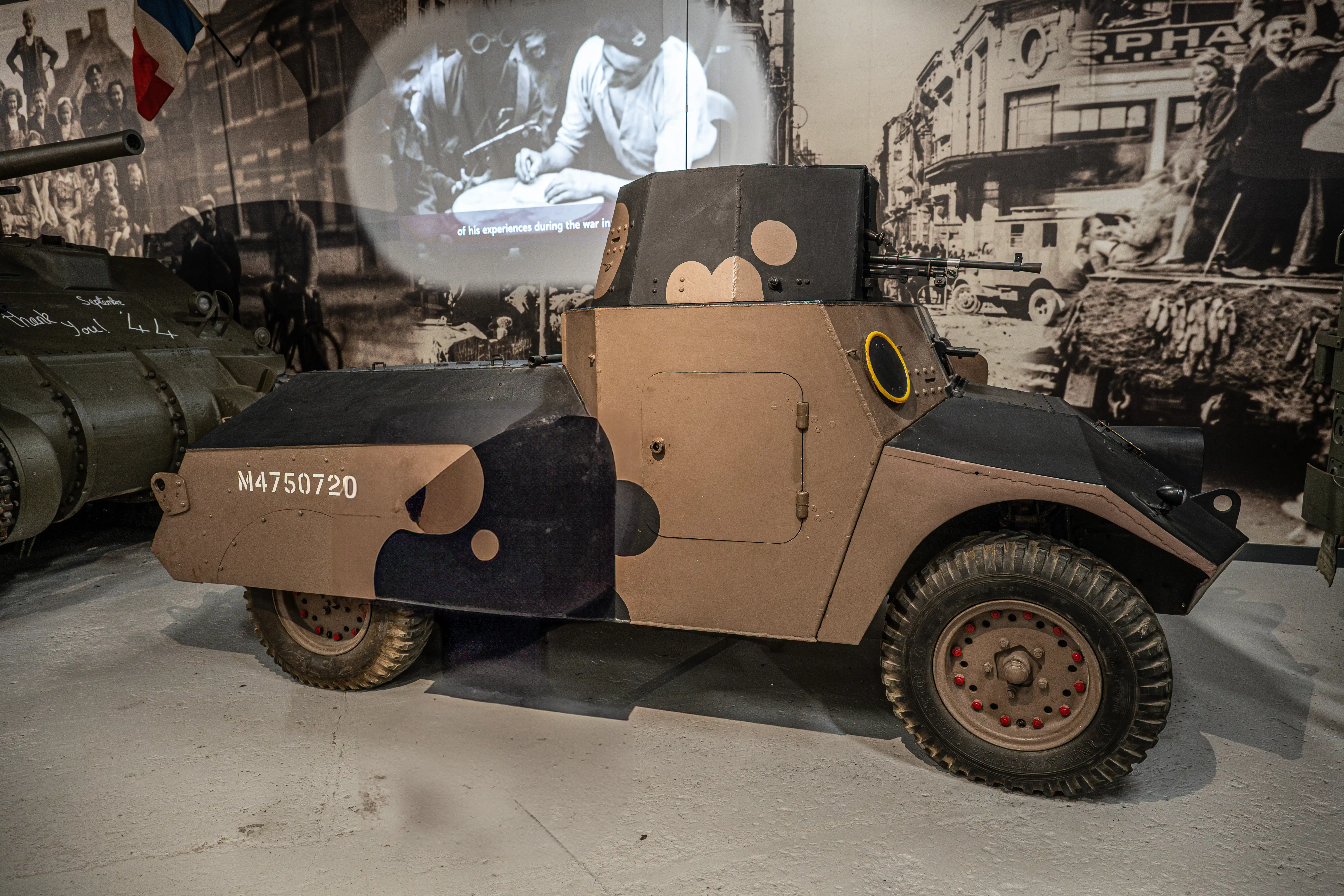 The image depicts a military armored vehicle, specifically an armored car, displayed in a museum setting. The vehicle has a camouflage paint scheme with a mix of brown and black colors. The side of the vehicle has a serial number 'M4750720' marked on it. The background features black-and-white photographs and a projection, likely depicting historical scenes or information related to the vehicle's use and context. The overall scene suggests an exhibit focused on historical military equipment, possibly from World War II.
