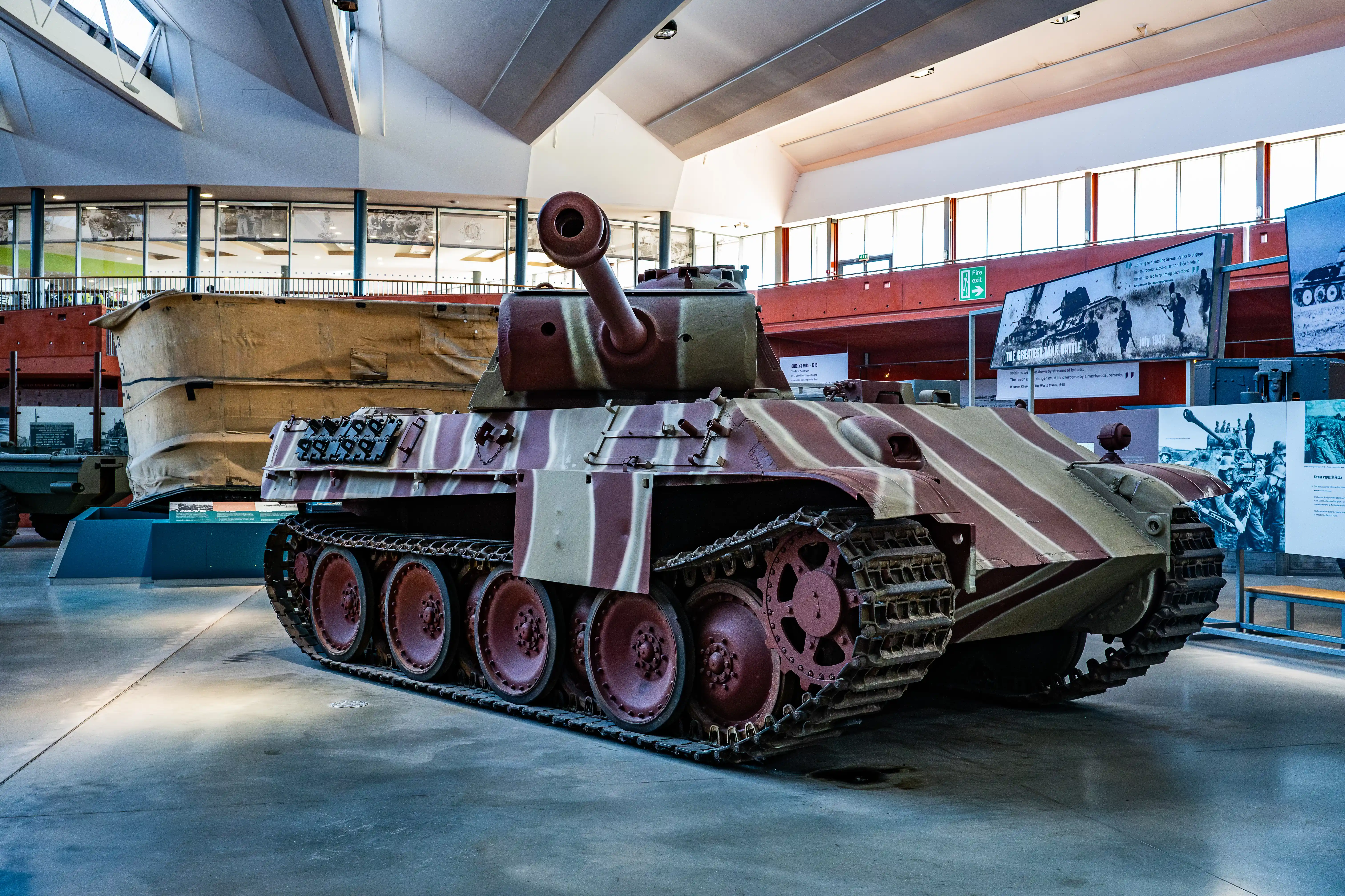 The image depicts a military tank displayed in a museum setting. The tank is painted in a camouflage pattern of red and white stripes. It is positioned on a smooth floor with large wheels and tracks designed for rugged terrain. In the background, there are informational displays and another large military vehicle, possibly a boat or a submarine, covered with a tarp. The museum has a modern design with large windows allowing natural light to illuminate the exhibits.