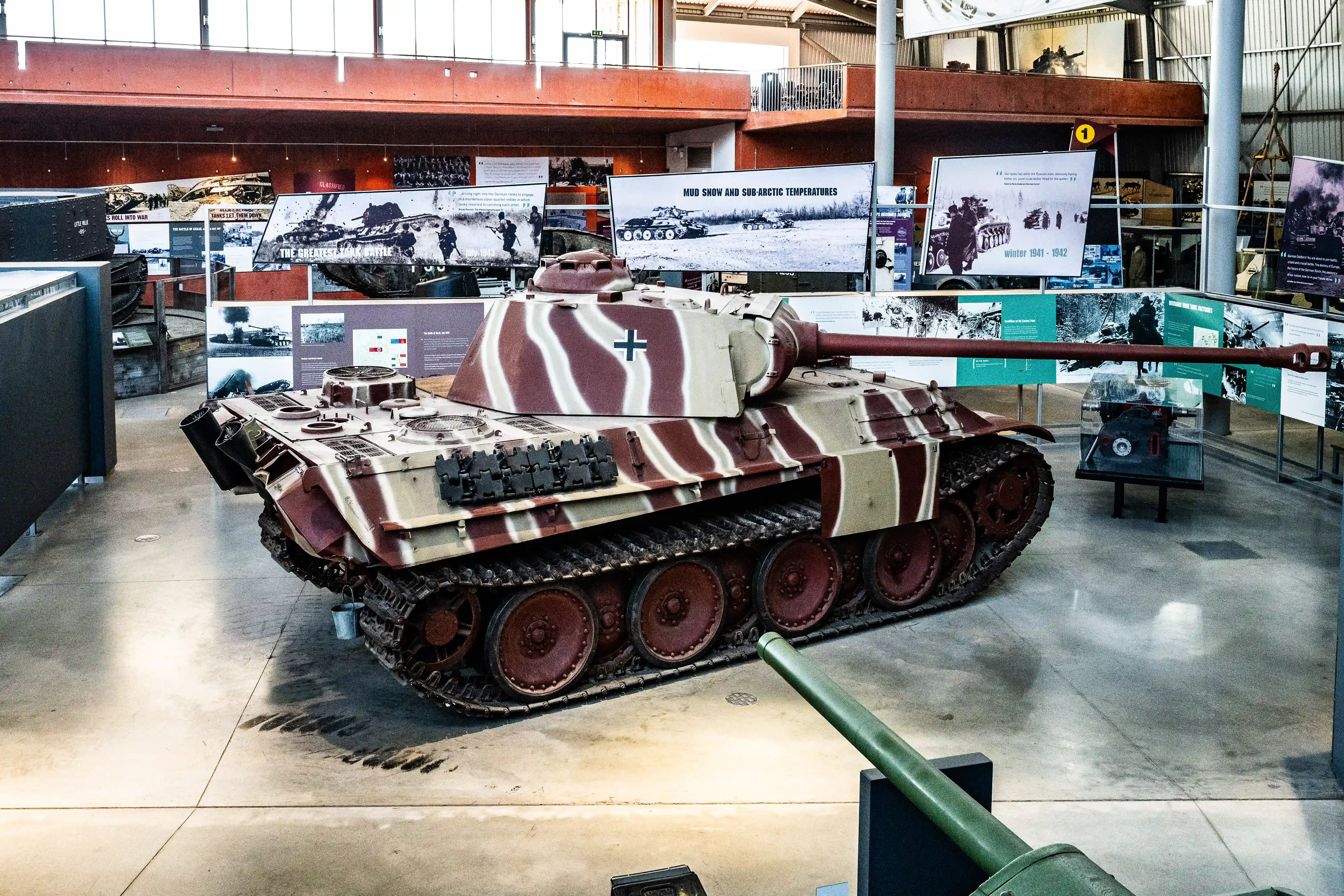 The image depicts a military history museum exhibit featuring a large, camouflage-painted tank, likely from World War II, based on the surrounding informational displays and photographs. The exhibit includes various panels detailing historical contexts, such as 'Mud, Snow, and Sub-Arctic Temperatures,' and 'Winter 1941-1942,' suggesting the tank's operational environments and historical significance. The museum setting is modern, with a spacious, well-lit interior and multiple floors of exhibits.