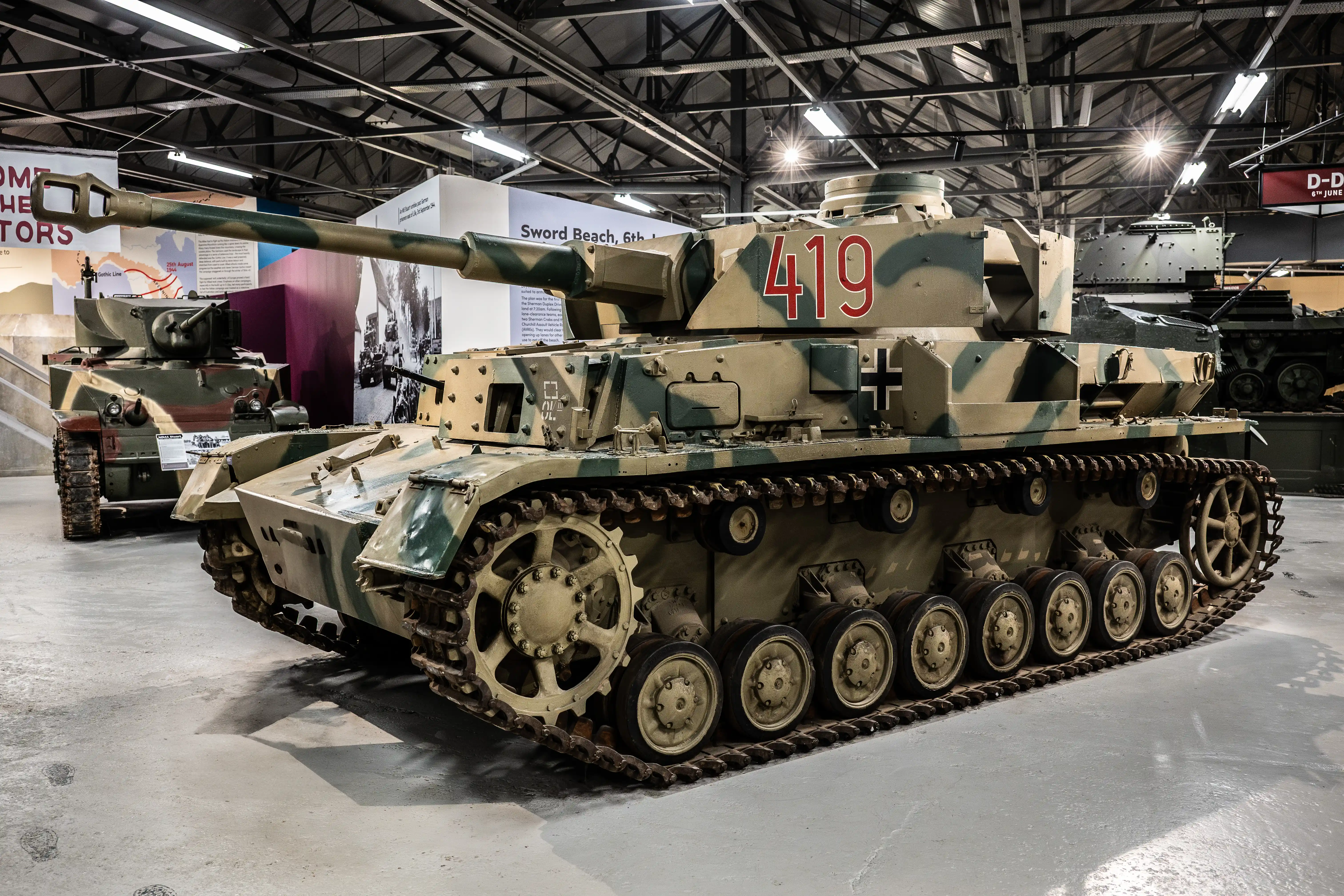 The image shows a museum exhibit featuring a World War II-era German Panzerkampfwagen VI Tiger Ausf E tank, identified by the number 419 on its turret. The tank is displayed indoors, with camouflage paint and a prominent Iron Cross marking. Surrounding displays and other tanks are visible in the background, suggesting a focus on historical military equipment and the events of D-Day.