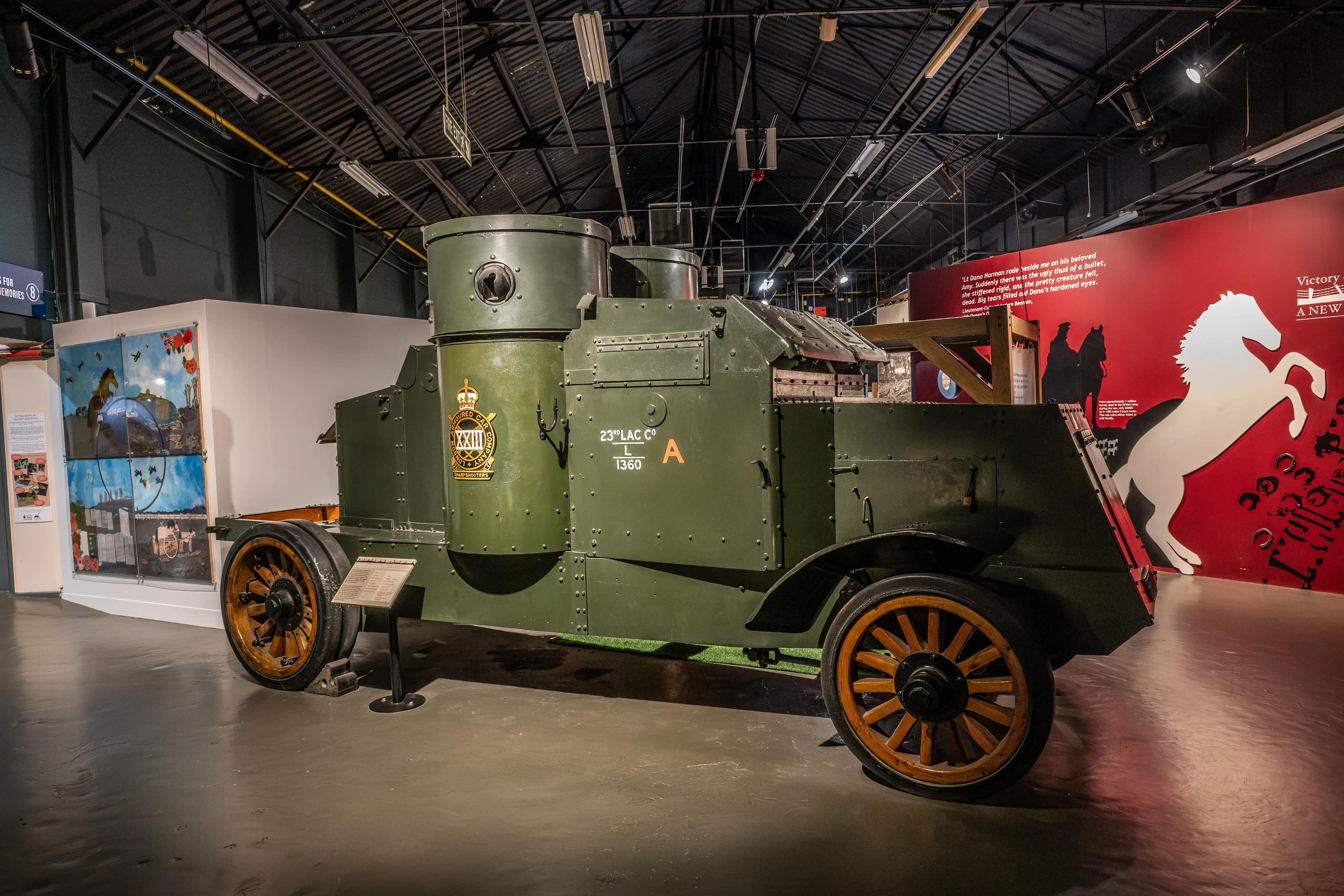 The image depicts a vintage military vehicle, specifically an armored car, displayed in a museum setting. The vehicle is painted in a dark green color and features wooden-spoked wheels. It has various markings and insignia, including the text '23rd LAC CO' and 'L 1360' on its side. The museum exhibit includes informational displays and a large red backdrop with a white horse illustration, suggesting a historical or military theme.