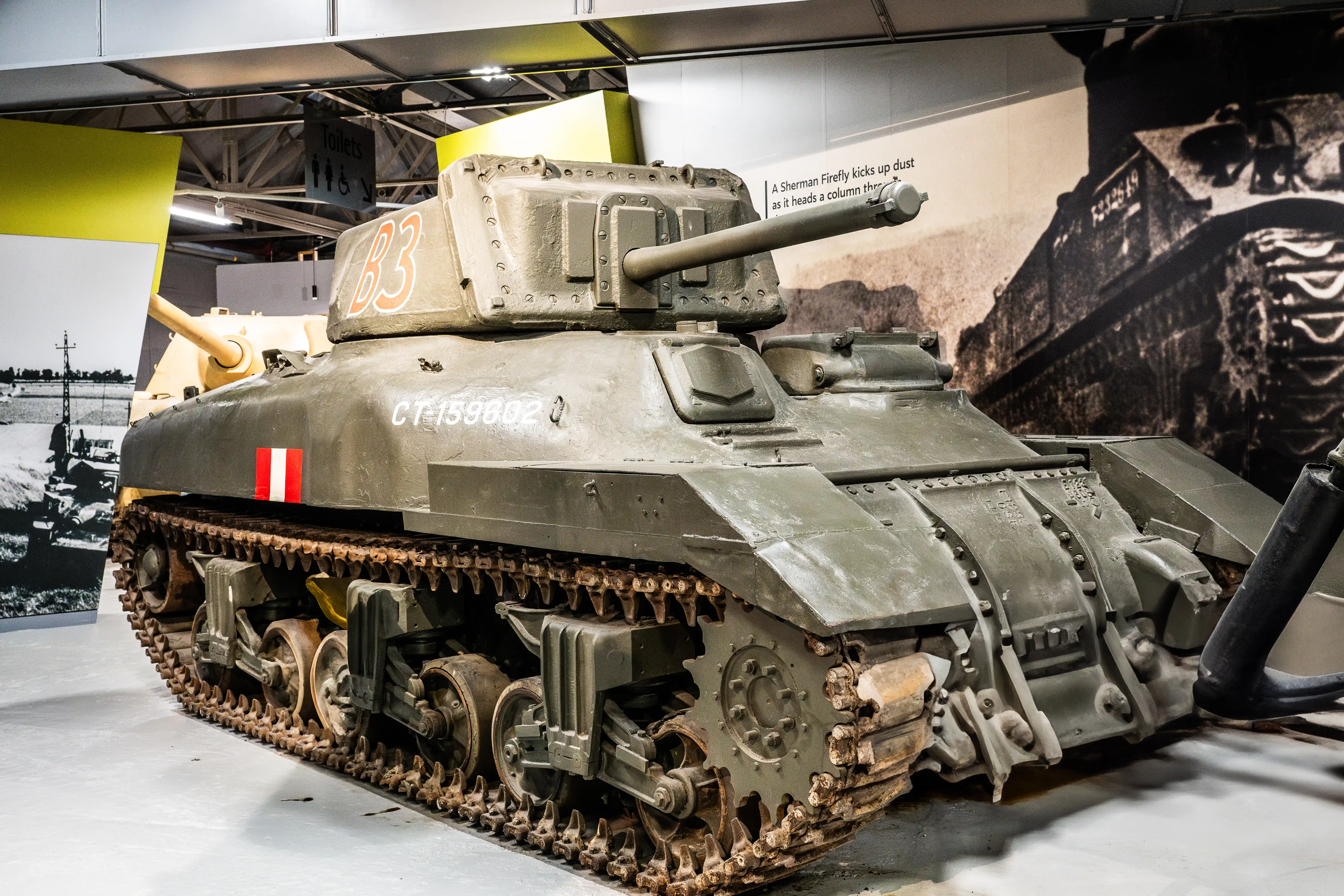 The image depicts a Sherman Firefly tank, a World War II-era tank known for its powerful 17-pounder gun. The tank is displayed in a museum setting, showcasing its detailed features and markings, including the identification number '23' and 'CT 159002'. The background includes historical photographs and text, providing context about the tank's use during the war.