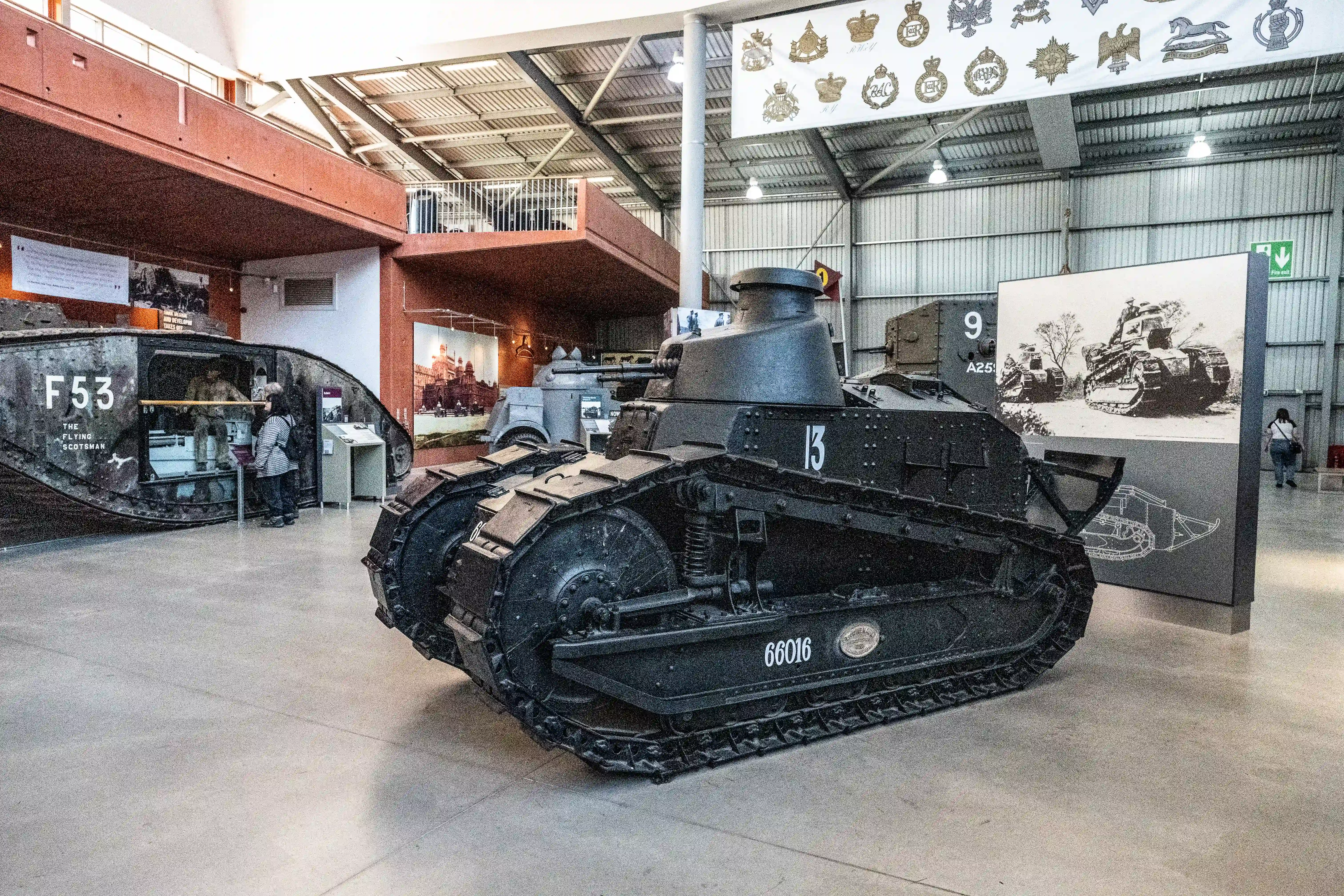 The image depicts a museum exhibit featuring historical military tanks. The central focus is on a black tank with the number 13 prominently displayed on its side. Other tanks and military artifacts are also visible in the background, along with informational displays and photographs. The museum setting includes a high ceiling, various insignia on the walls, and a few visitors observing the exhibits.