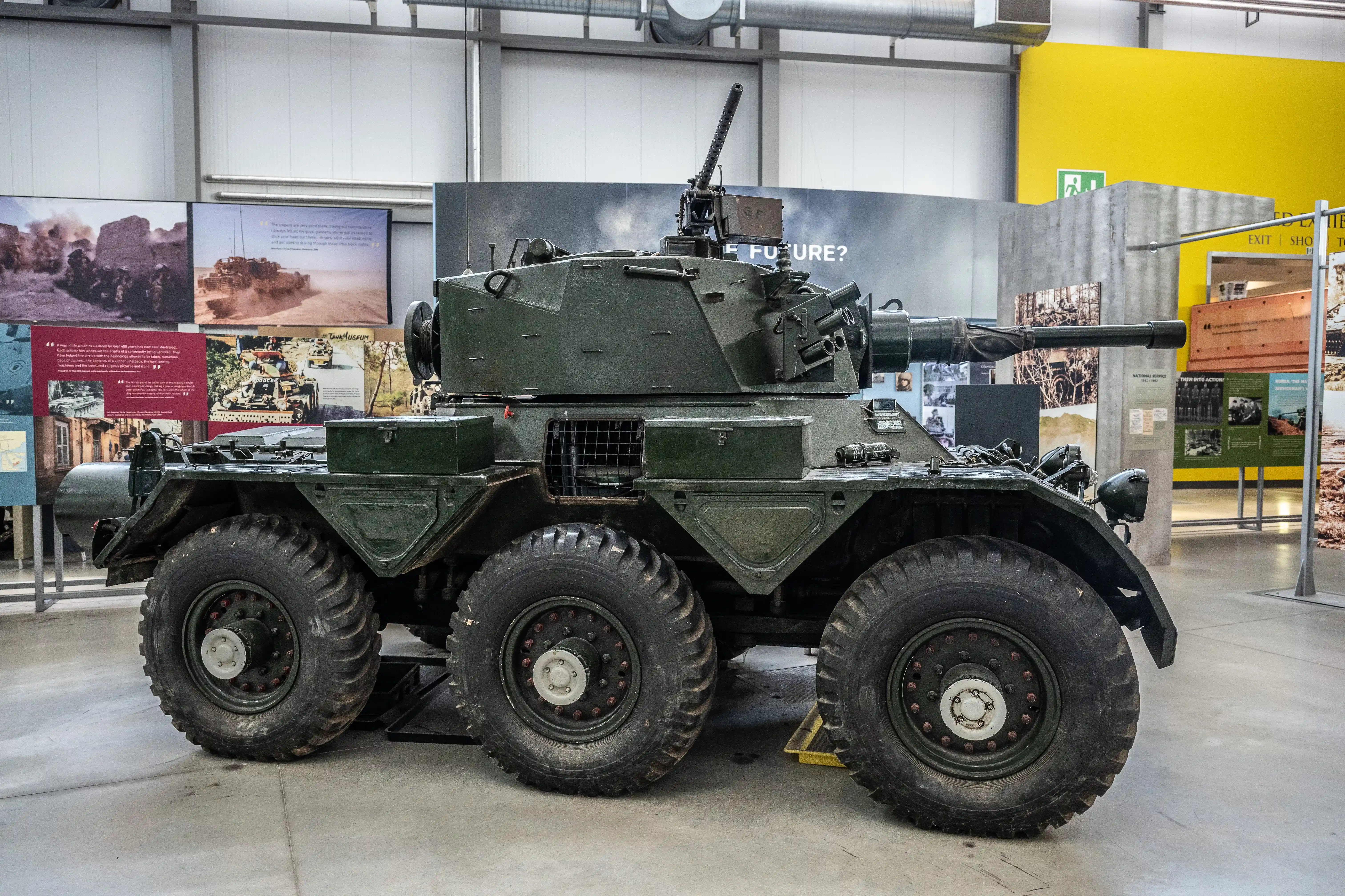 The image depicts a military vehicle on display in a museum or exhibition setting. The vehicle is a six-wheeled armored car equipped with a large gun turret on top. The surroundings include informational panels and displays, suggesting that the vehicle is part of an exhibit on military history or technology. The setting appears to be indoors, with a clean and organized environment. The vehicle's design and the context of the display indicate it may be a historical or prototype model.