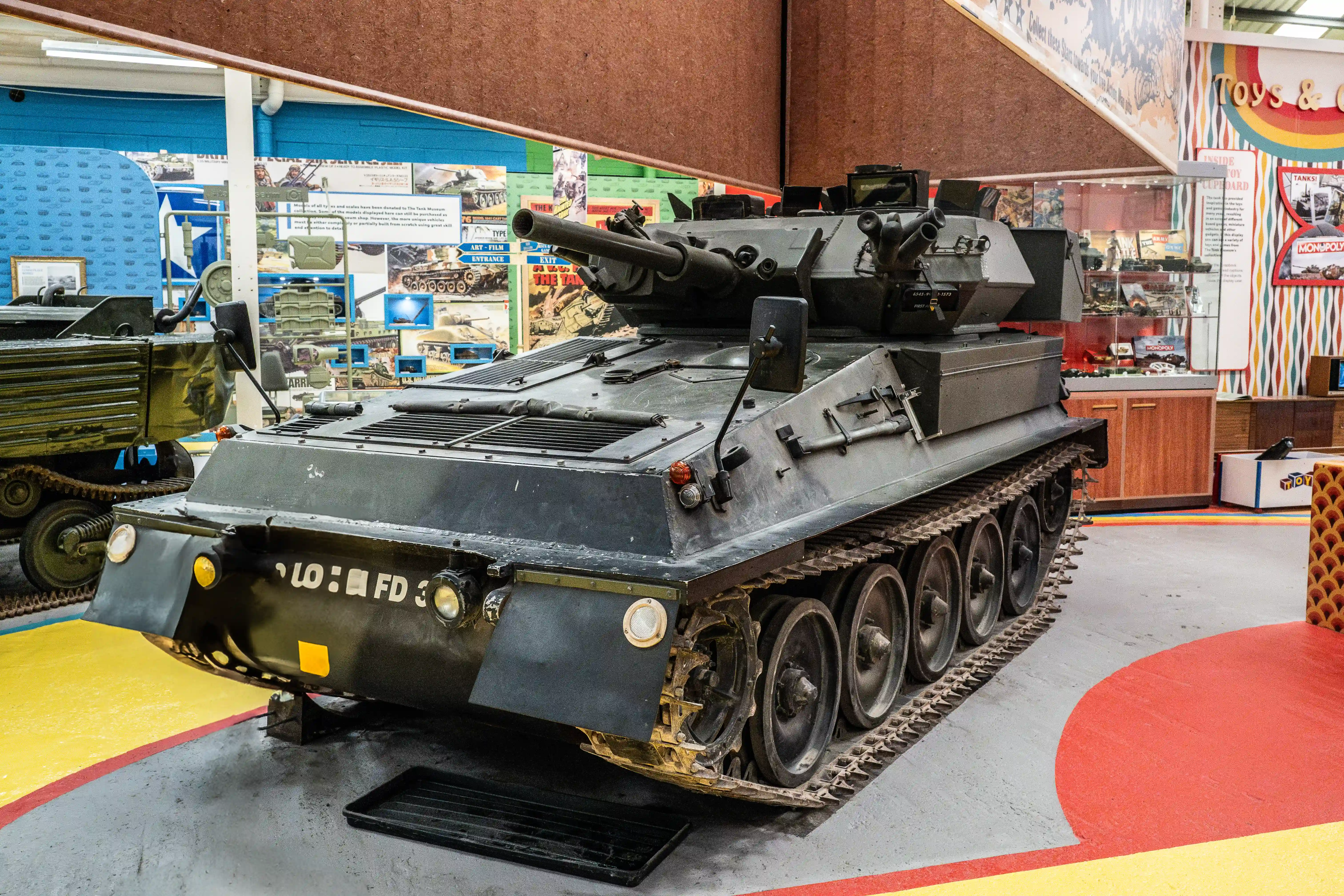 The image shows a military tank on display in a museum setting. The tank is positioned on a colorful floor with a yellow, red, and blue design. The tank has a large gun mounted on top and is surrounded by various informational posters and displays related to military vehicles. The environment suggests an educational exhibit aimed at informing visitors about the history and types of tanks.