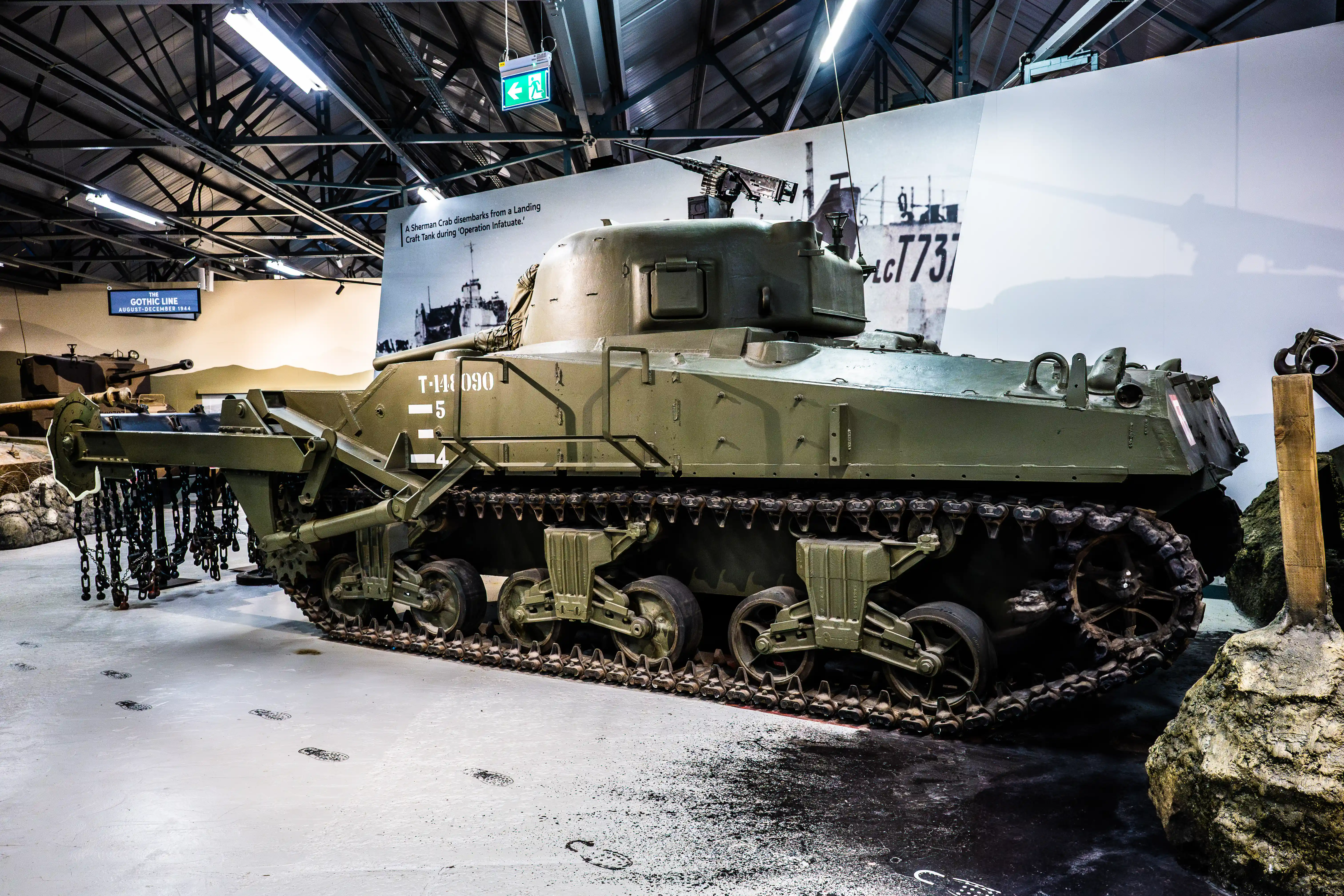 The image depicts a military tank, specifically a Sherman Crab, displayed in a museum setting. The tank is equipped with a flail system used for clearing landmines, as indicated by the chains and rollers attached to its front. The tank is marked with the identification number T-144090 and the number 5. The background features a large photograph of a similar tank disembarking from a landing craft during Operation Infatuate, which took place along the Gothic Line from August to December 1944. The setting appears to be an indoor exhibit, with a high ceiling and artificial lighting.