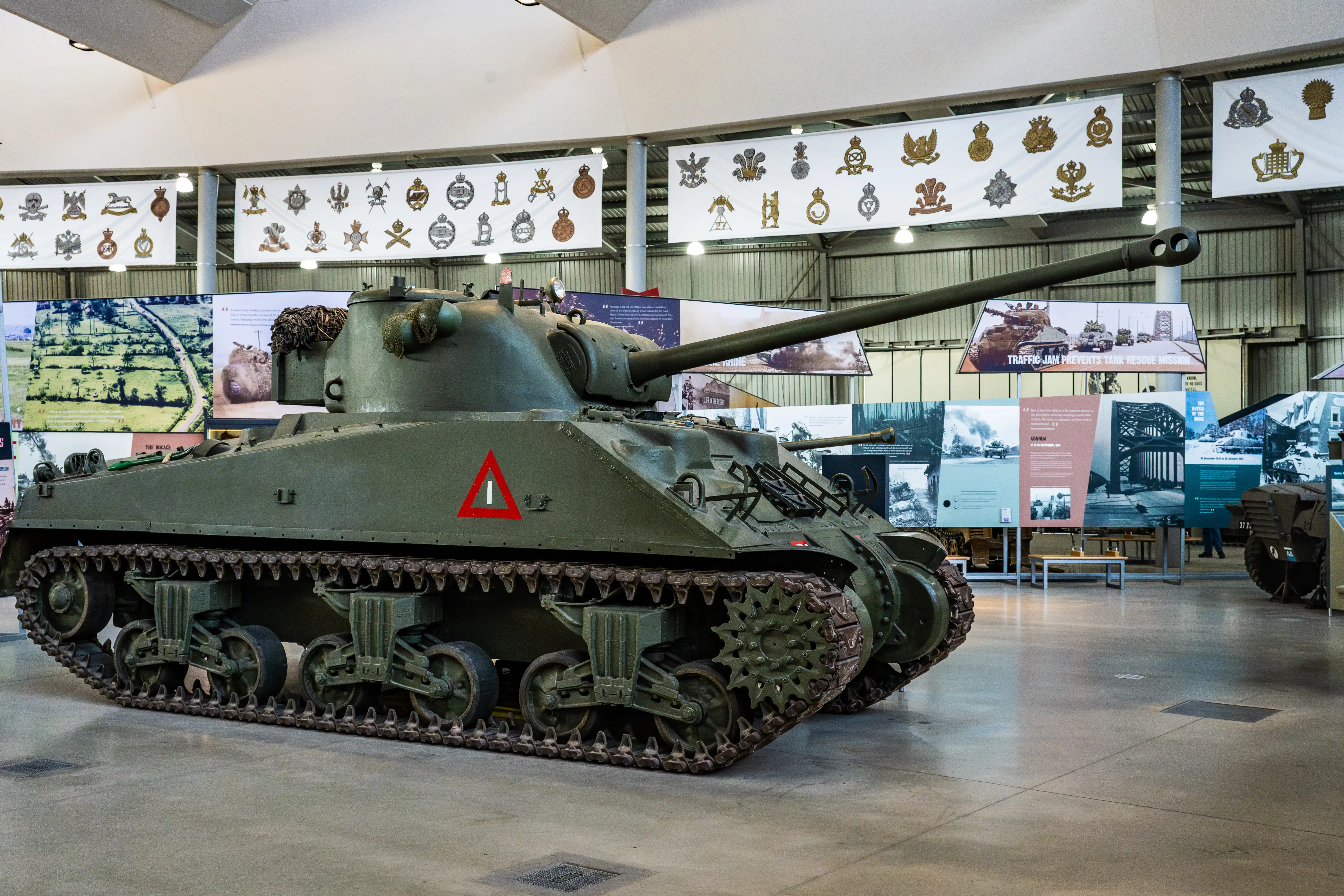 The image depicts a military museum exhibit featuring a large, green, tracked armored vehicle, specifically a tank, prominently displayed in the center. The tank has a long barrel extending from its turret and a red triangle marking on its side. Surrounding the tank are various informational displays and banners showcasing historical photographs, text, and insignia related to military history. The setting appears to be an indoor exhibit space with a high ceiling and ample lighting.