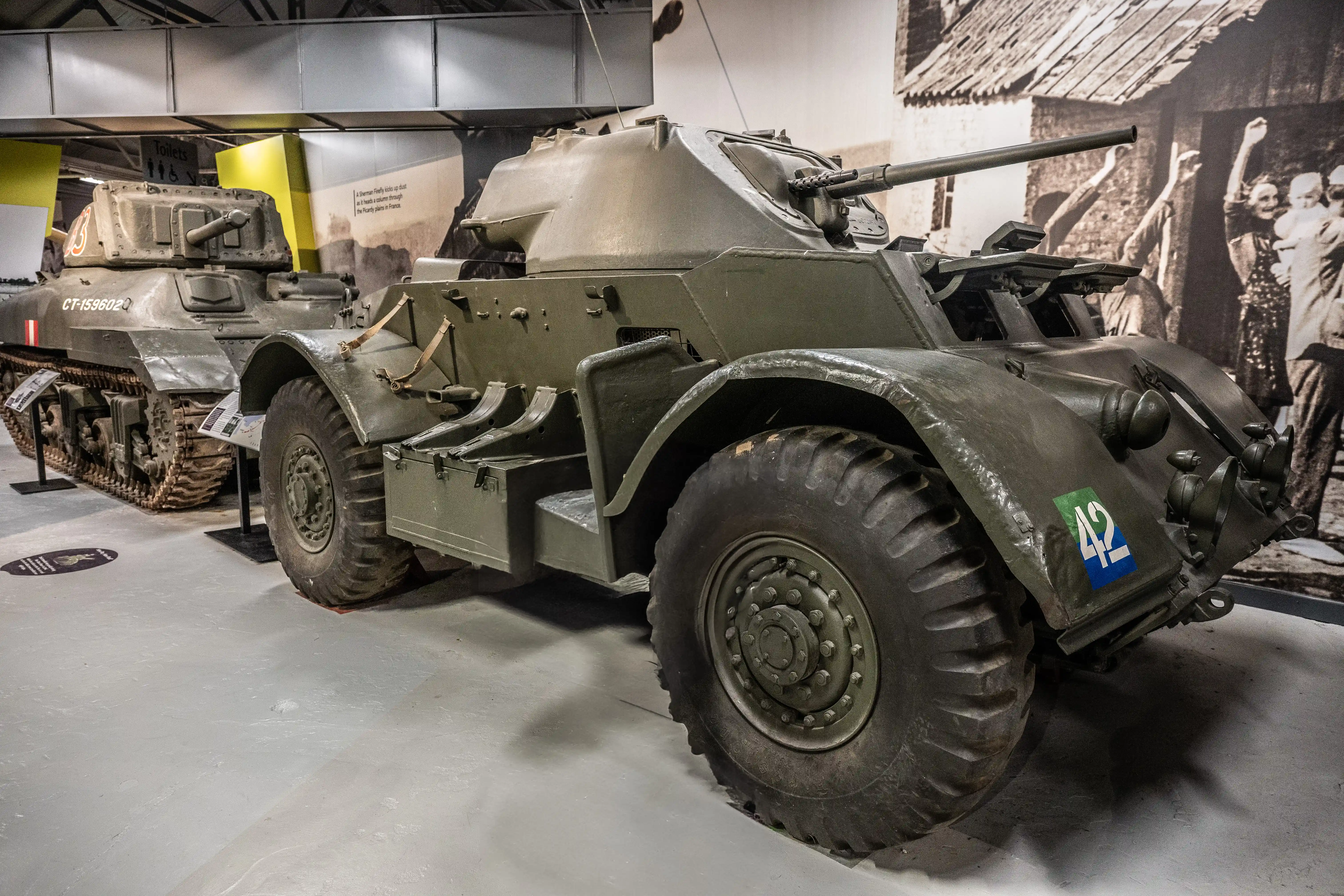 The image shows a military vehicle, specifically an armored car, displayed in a museum setting. The vehicle has large, rugged tires and a mounted gun on top. It is painted in a military green color and features various markings and equipment. The background includes other military vehicles and historical photographs, suggesting the exhibit focuses on military history.