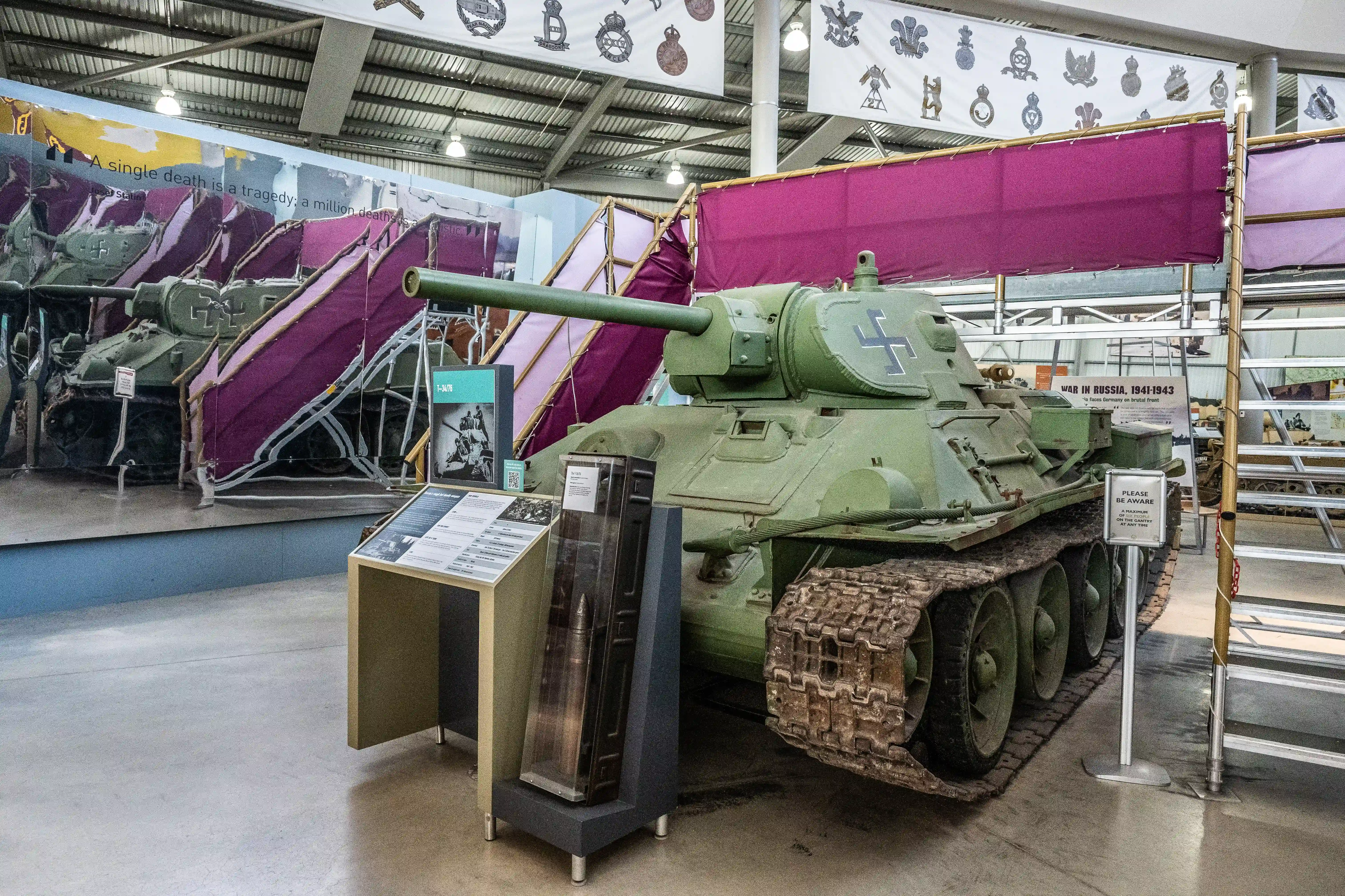 The image depicts a military museum exhibit featuring a green Soviet-era T-34 tank with a white cross symbol on its side. The tank is displayed indoors on a concrete floor with informational placards in front of it, providing context about its historical significance. The exhibit is part of a larger display area with various military artifacts and banners overhead showcasing different military insignias. The setting appears to be well-lit and organized, likely aimed at educating visitors about military history, specifically focusing on the period of the war in Russia from 1941 to 1943.
