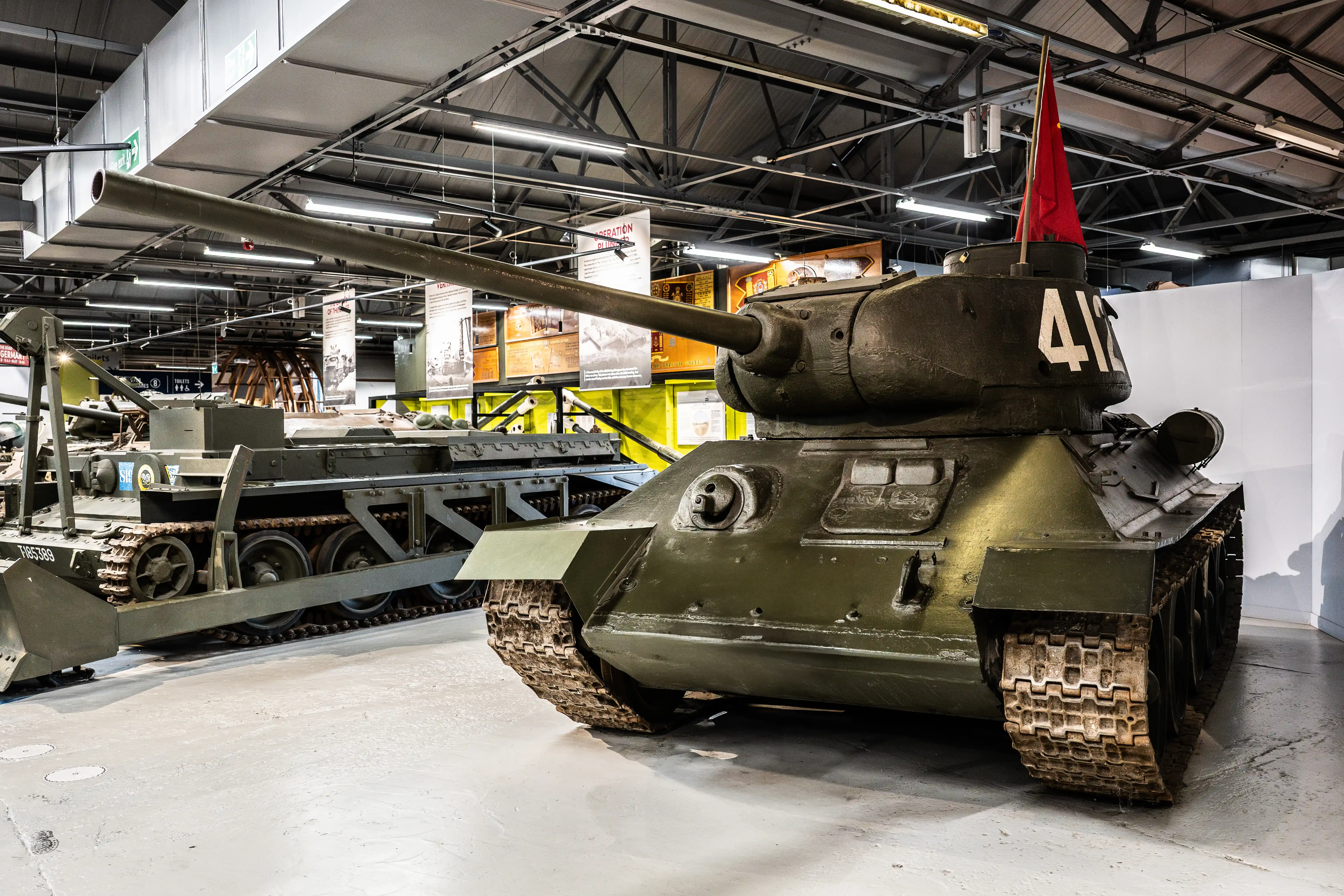 The image depicts a military museum exhibit featuring a Soviet T-34 medium tank, prominently marked with a red flag and the number 4 on its turret. The tank is displayed indoors, with various informational posters and other military equipment visible in the background. The setting appears to be well-lit and organized, likely part of a larger collection of historical military vehicles and artifacts.