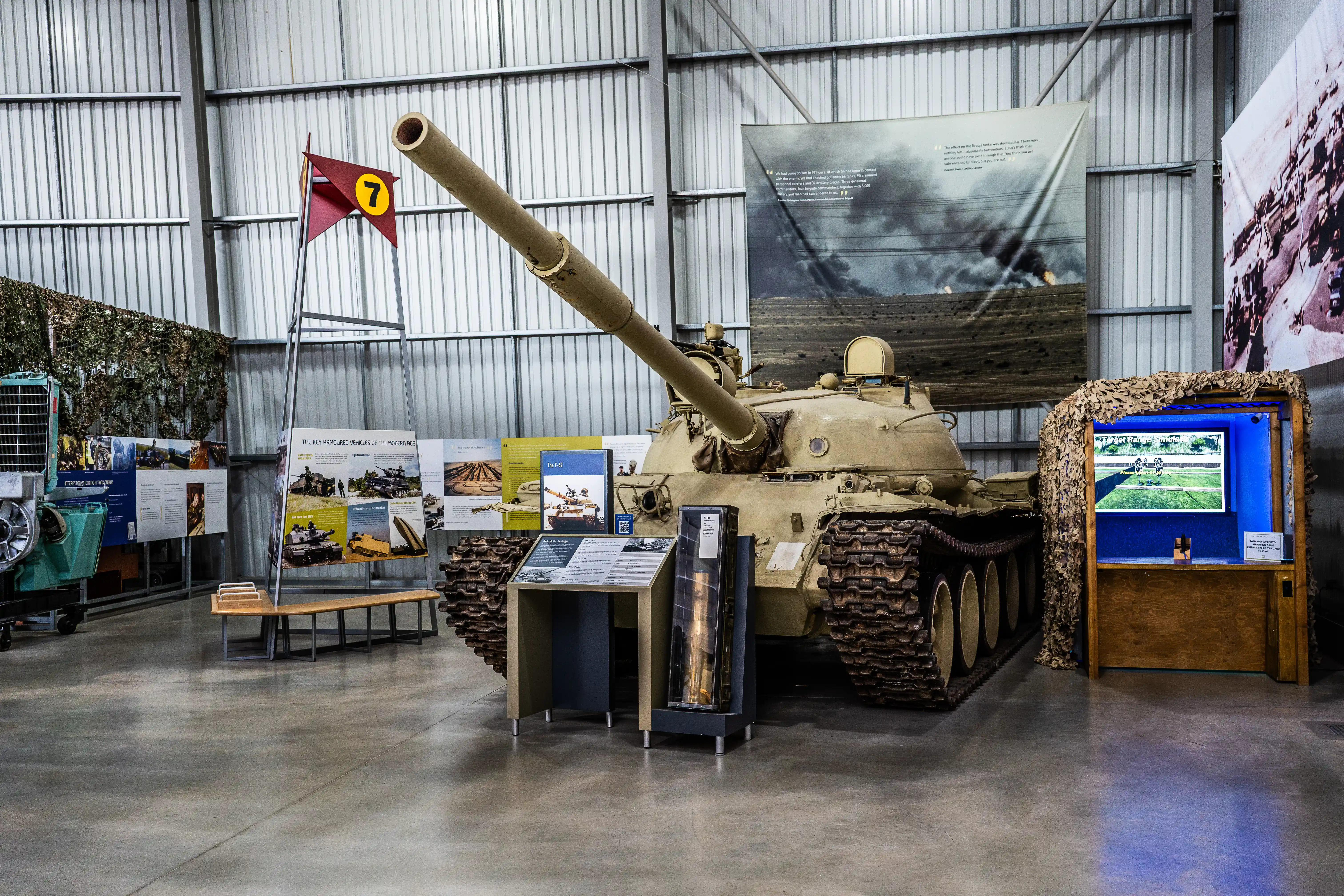 The image depicts an indoor exhibit featuring a military tank as the central piece. Surrounding the tank are various informational displays and panels providing context and details about the tank and related military vehicles. The exhibit is housed in a spacious, well-lit hangar-like structure with high ceilings and large windows. There are additional artifacts and educational materials placed around the tank, enhancing the educational experience for visitors. The overall setting suggests a museum or educational facility focused on military history and technology.