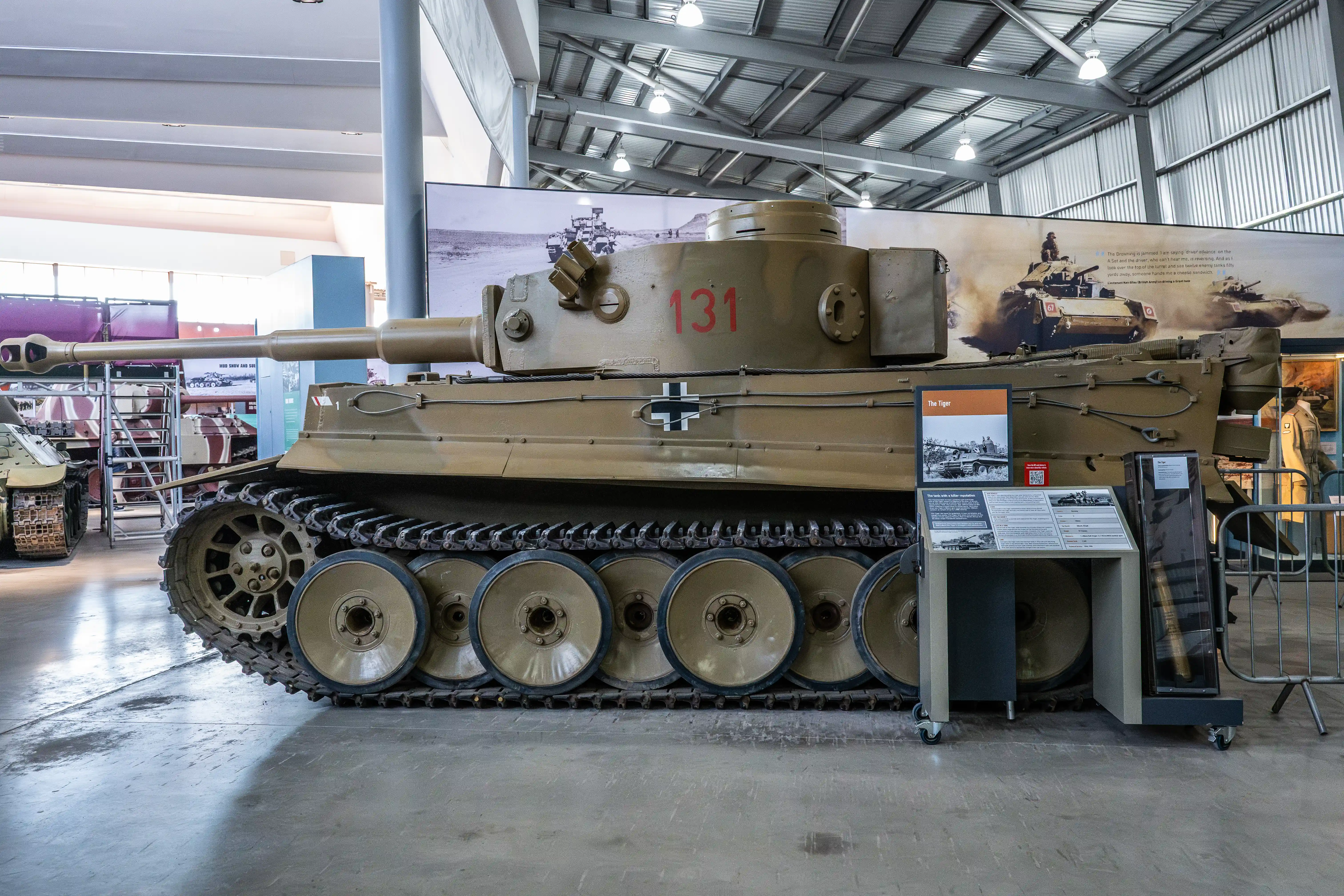 The image depicts a Tiger I tank on display in a museum. The tank, marked with the number 131, is a historical military vehicle known for its role in World War II. The exhibit includes informational placards providing context and details about the Tiger I tank, highlighting its significance and historical background.