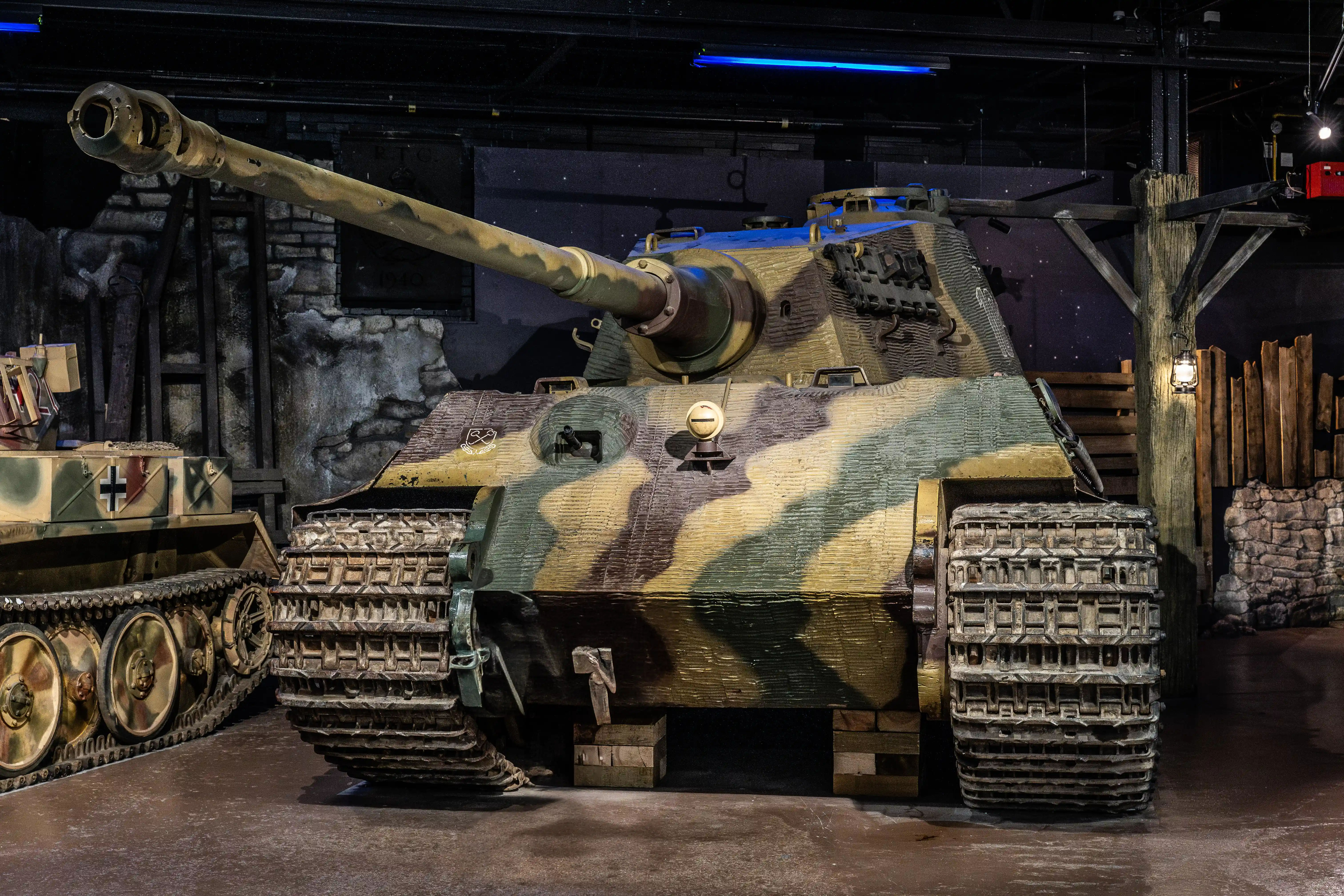 The image depicts a military tank, specifically a German Panther tank from World War II, displayed in a museum or exhibition setting. The tank is painted in a camouflage pattern and is positioned indoors, surrounded by a themed backdrop that includes brick walls and wooden supports. The tank features a prominent main gun and intricate details on its tracks and armor