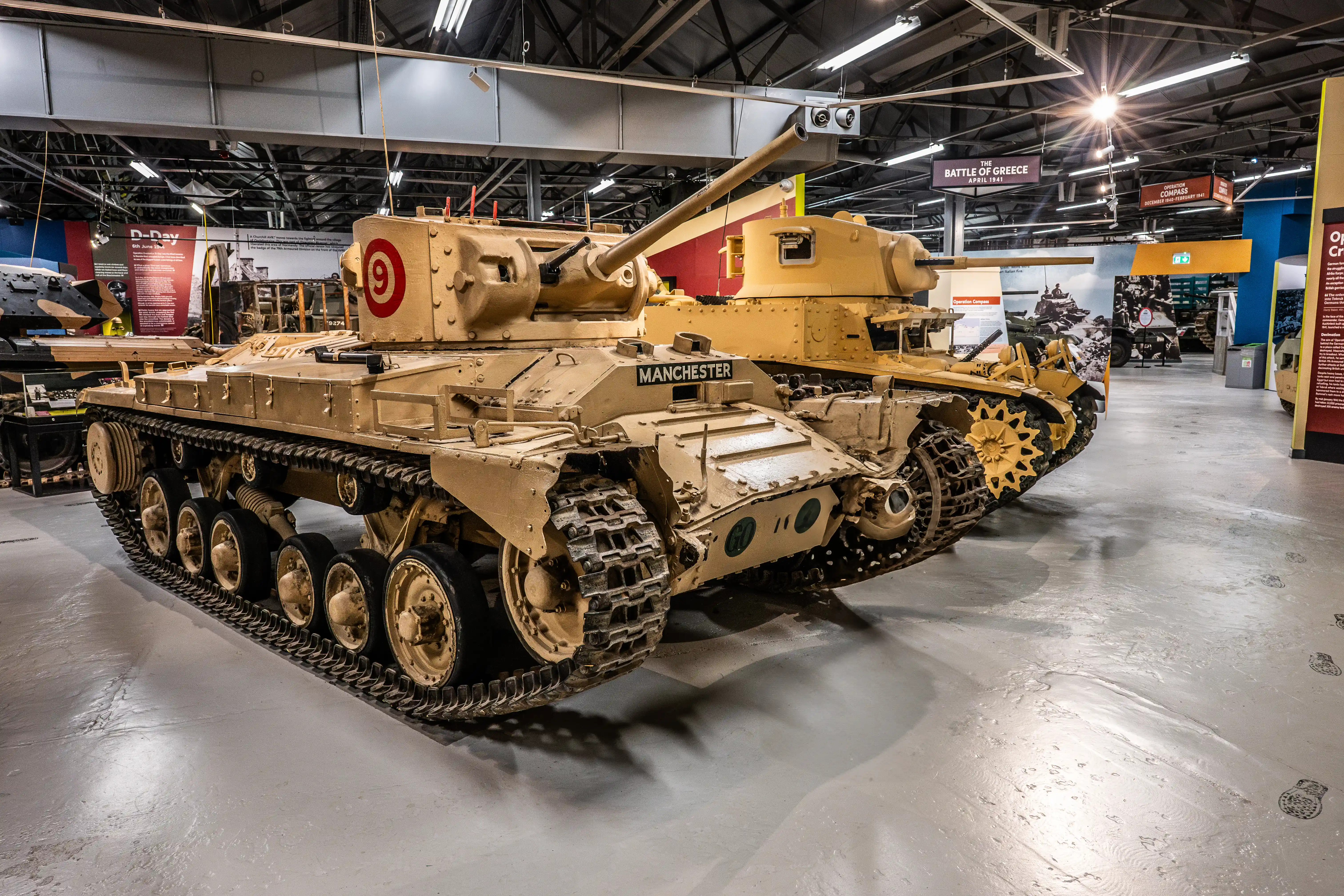 The image depicts a military museum exhibit featuring a tank named 'Manchester' with the number 9 marked on it. The tank is displayed indoors, with various informational panels and other military vehicles visible in the background.