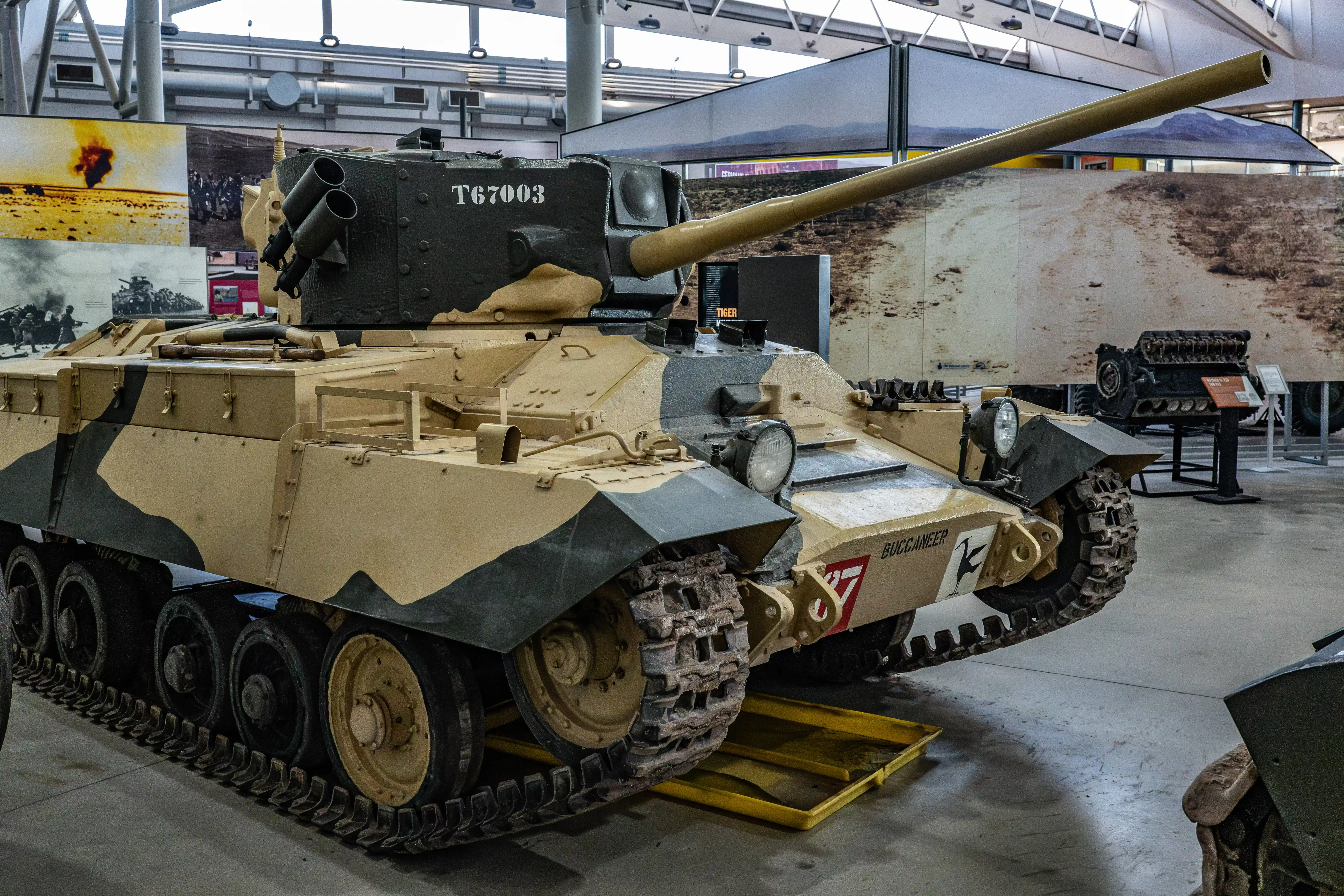 The image shows a military tank on display in a museum. The tank, identified as T67003 and named BUCCANEER, is a historical piece likely from World War II. It features a large gun barrel, camouflage paint, and various markings. The display area includes informational posters and other related exhibits, providing educational context about the tank and its historical significance.