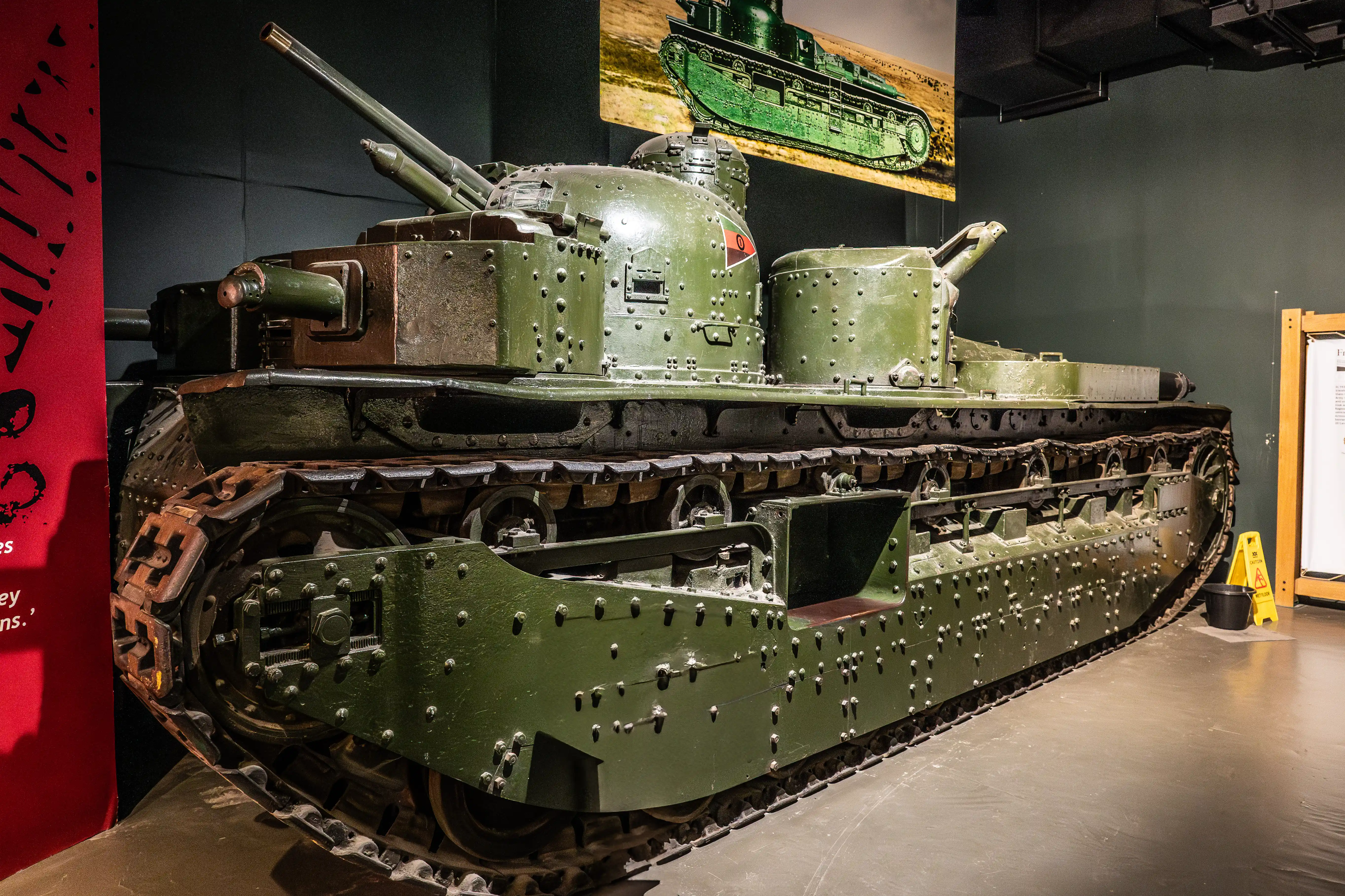 The image shows a historical military tank on display, likely in a museum setting. The tank is painted in a green color and has a riveted armor design. It features a turret with a mounted gun and various other armaments. The tank tracks and undercarriage are visible, showcasing the detailed mechanics of the vehicle. The background includes informational displays and a large poster of the tank.