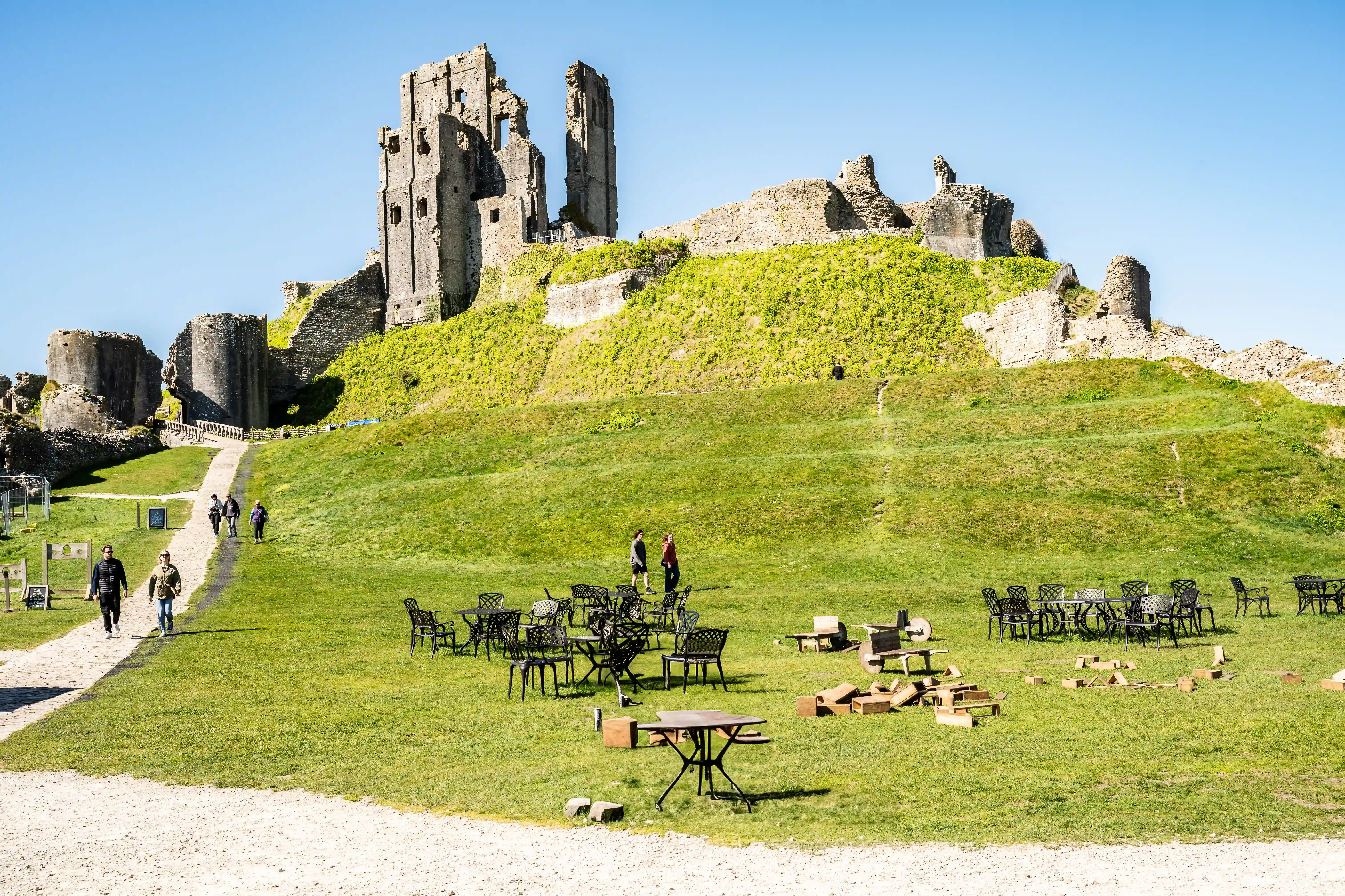 The image depicts the ruins of a castle on a grassy hill, with several people walking on a path leading up to the ruins. In the foreground, there are numerous outdoor tables and chairs, some of which are stacked or in disarray, suggesting a temporary or seasonal outdoor seating area, possibly for a café or restaurant. The scene is set on a clear, sunny day with a blue sky.