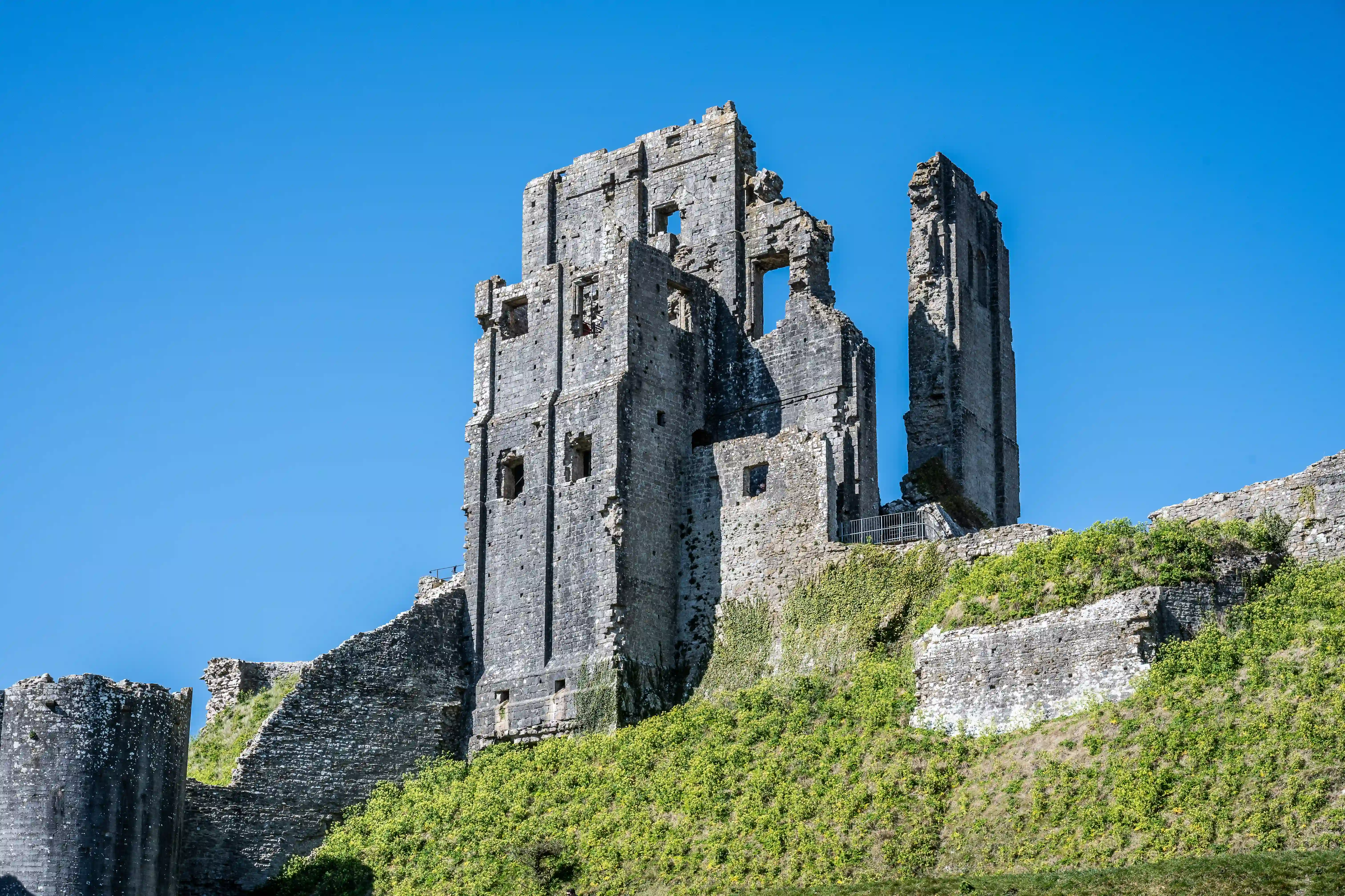 The image depicts the ruins of a medieval castle, likely located on a hillside. The structure is made of stone and shows significant signs of decay, with several walls and towers partially collapsed. The sky is clear and blue, indicating a sunny day. Vegetation, including grass and small shrubs, has grown around and over parts of the ruins, suggesting that the site has been abandoned for a considerable period. The overall scene conveys a sense of historical significance and the passage of time.