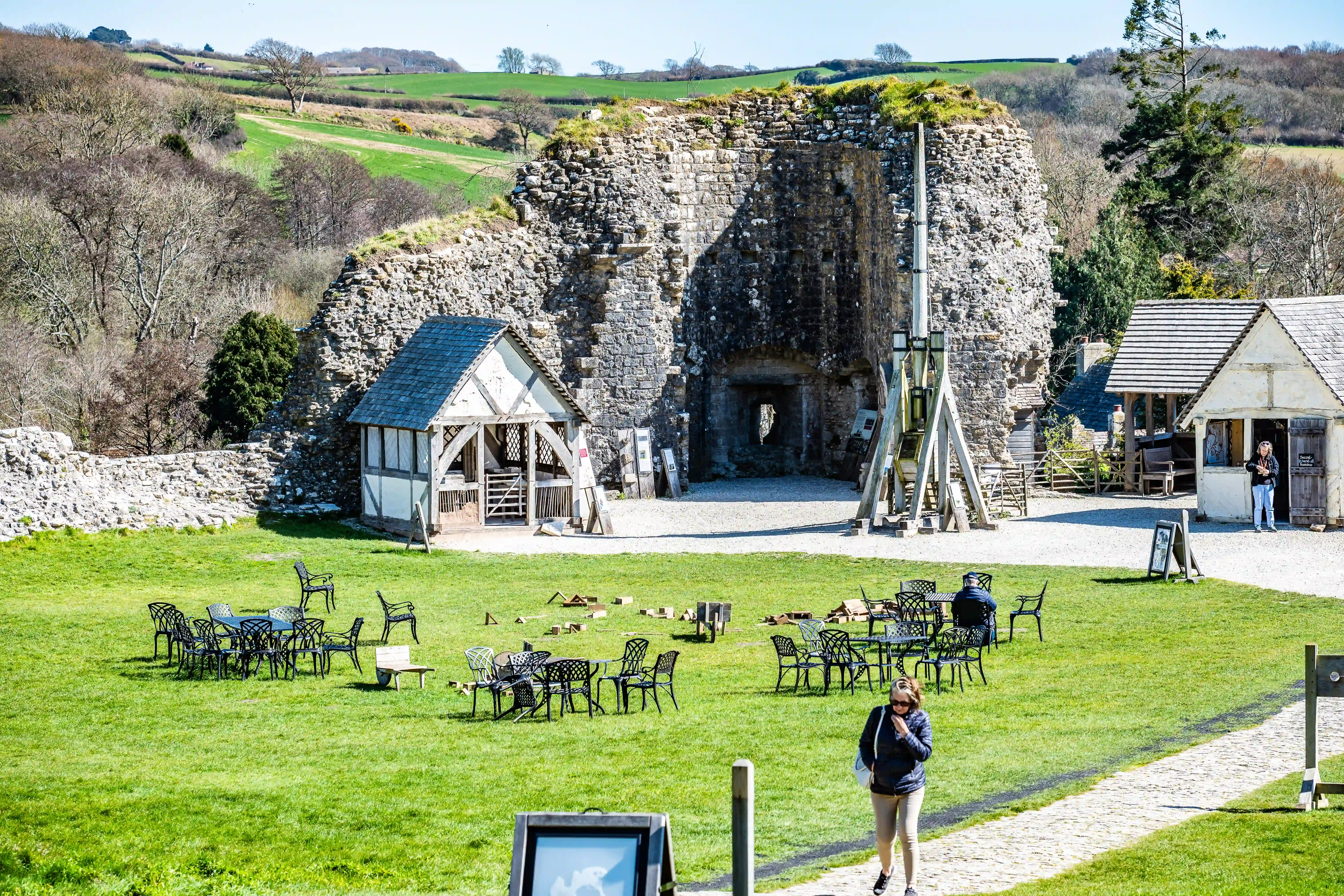 This image showcases the ruins of a historic castle set within a lush green landscape. Several small, rustic buildings, possibly for vendors or information, are situated within the castle grounds. In the foreground, neatly arranged metal tables and chairs suggest a designated area for visitors to relax. A trebuchet, a historical siege engine, stands prominently next to the main stone structure, adding to the historical atmosphere. People are visible enjoying the space, with one person walking on a stone path and others seated at tables. The background features rolling hills and trees under a clear blue sky.