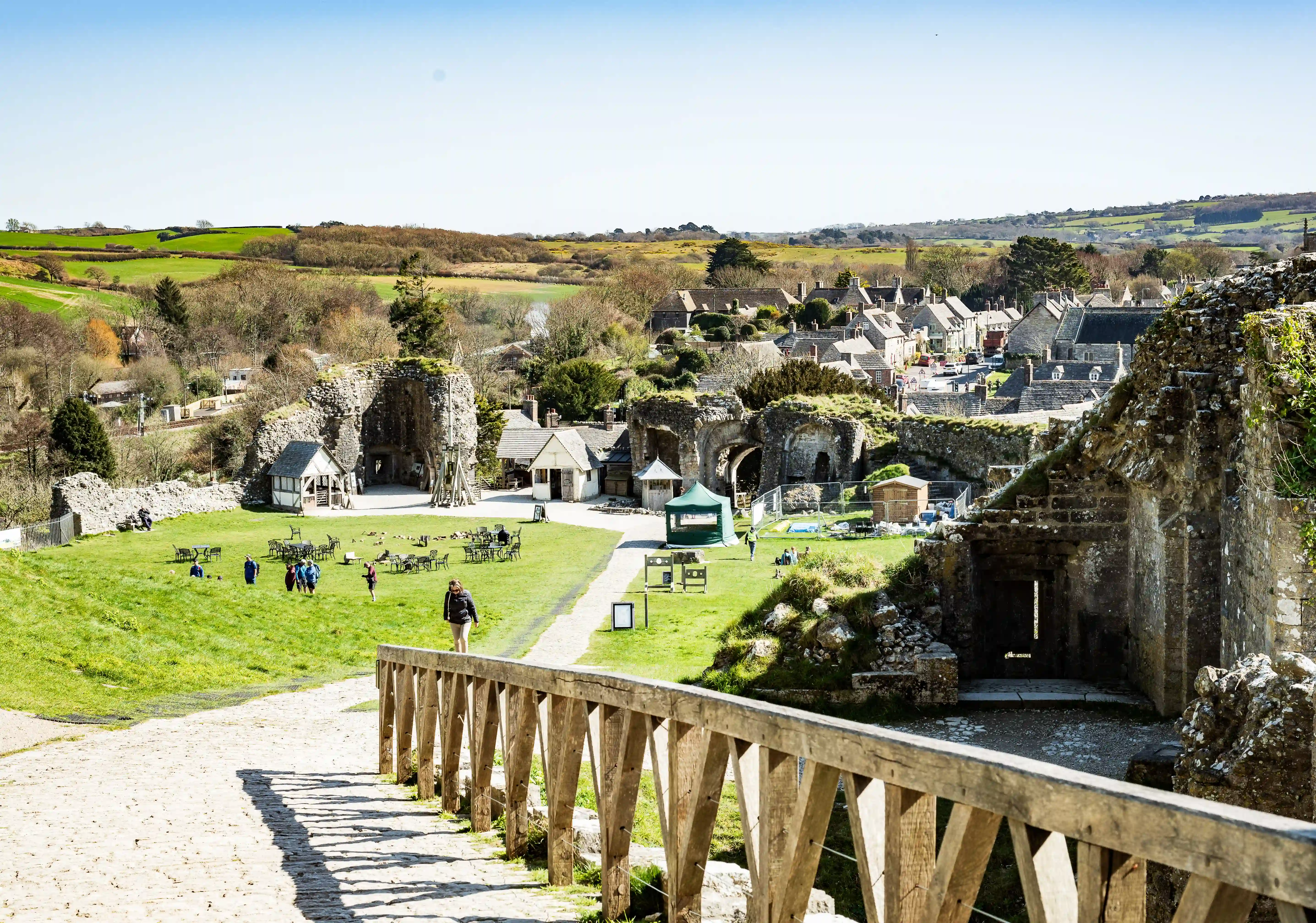 The image depicts a scenic view of a historic site with ancient stone structures, lush greenery, and a clear blue sky. The area features pathways, wooden railings, and a few small buildings, likely used for tourism or educational purposes. Visitors are seen walking and exploring the site, which is surrounded by rolling hills and a quaint village in the background.
