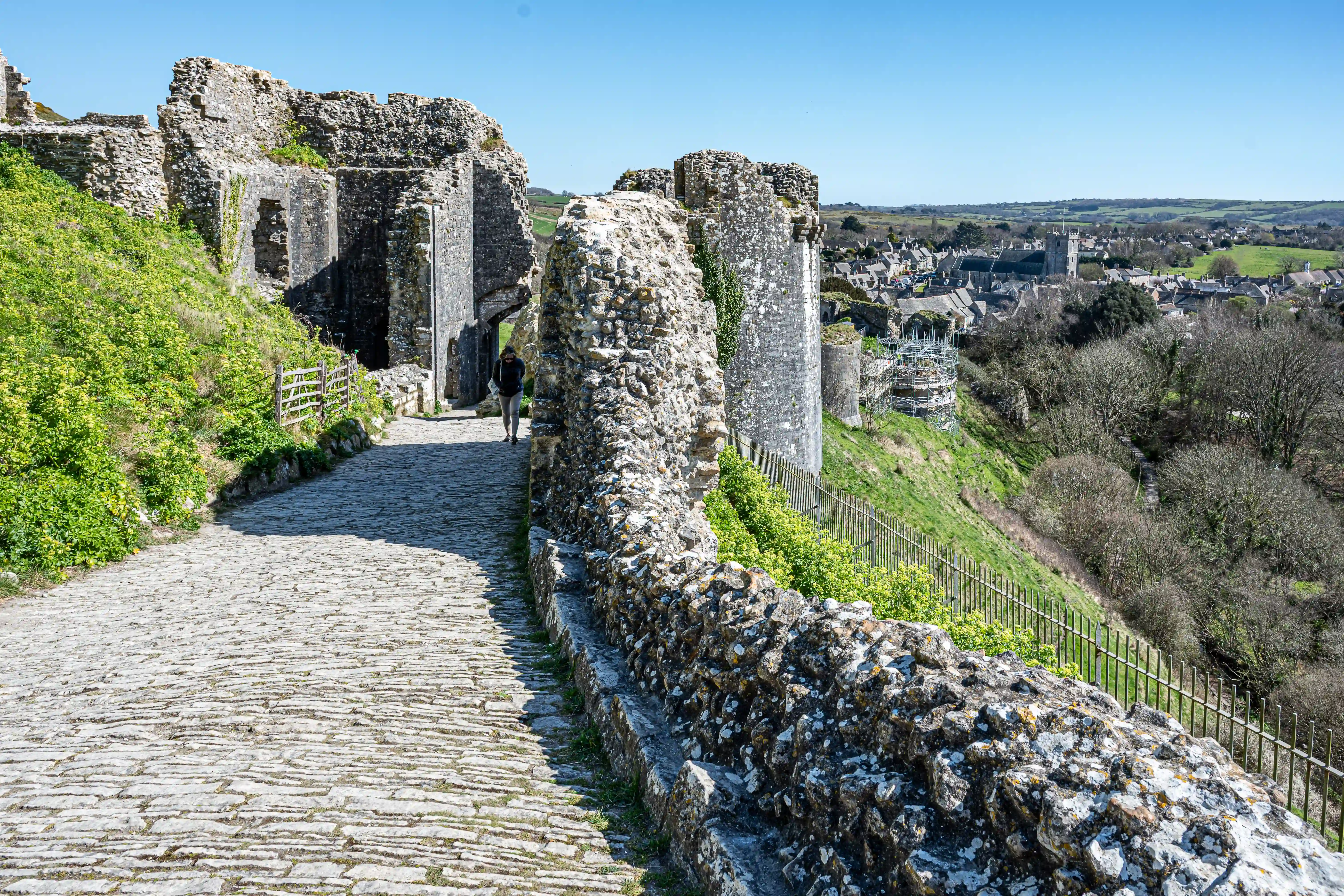 The image depicts the ruins of an old castle with a cobblestone path leading through the entrance. The castle walls are made of stone and partially overgrown with vegetation. A person is seen walking along the path, and the background shows a scenic view of a town with houses and a church, surrounded by green fields and hills under a clear blue sky.