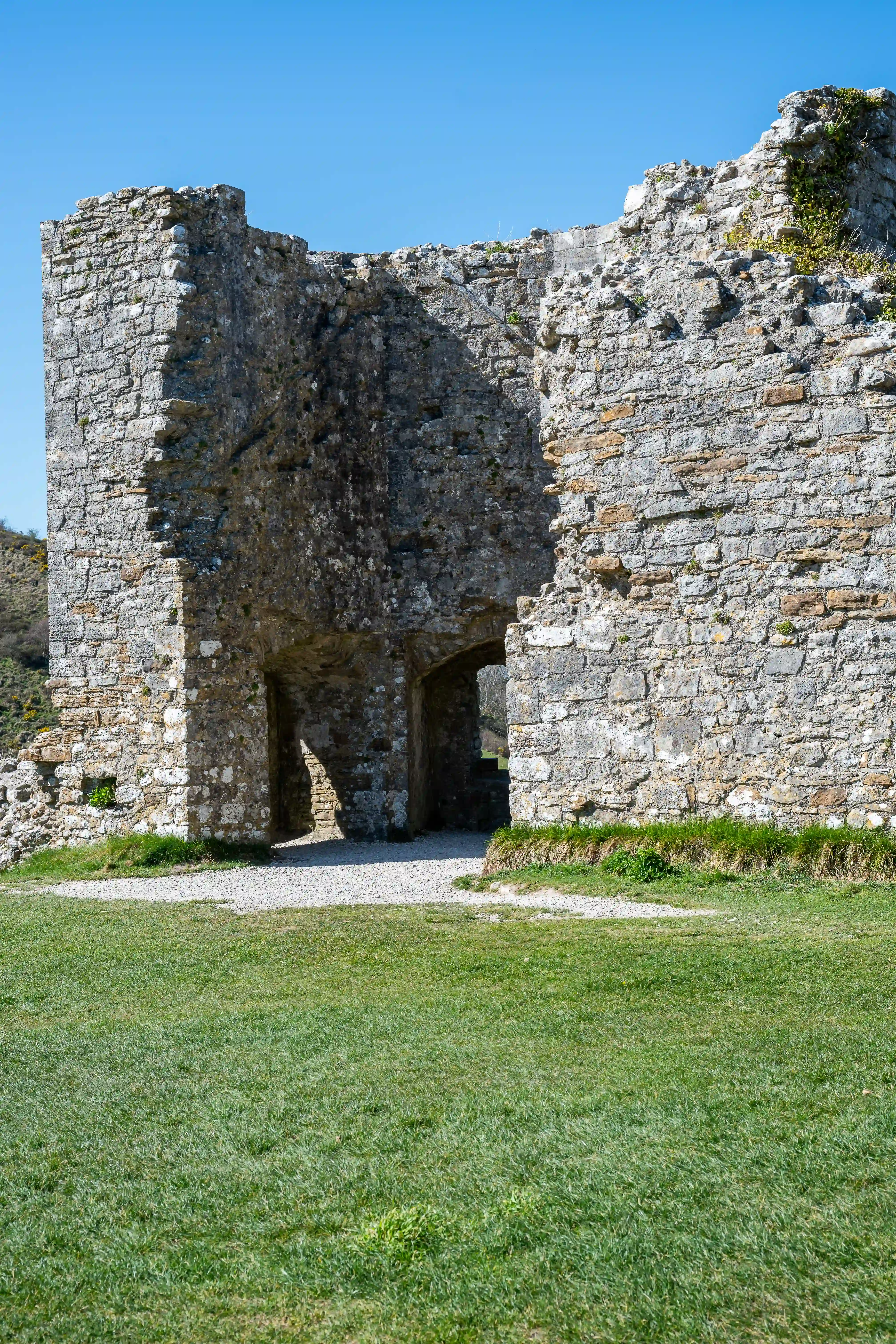 The image depicts the ruins of an ancient stone structure, likely a castle or fortification. The walls are made of large, irregularly shaped stones, showing significant wear and age. The structure features an arched entrance with a path leading through it, surrounded by a grassy area. The sky is clear and blue, indicating a sunny day. The overall scene suggests a historical site, possibly from medieval times.