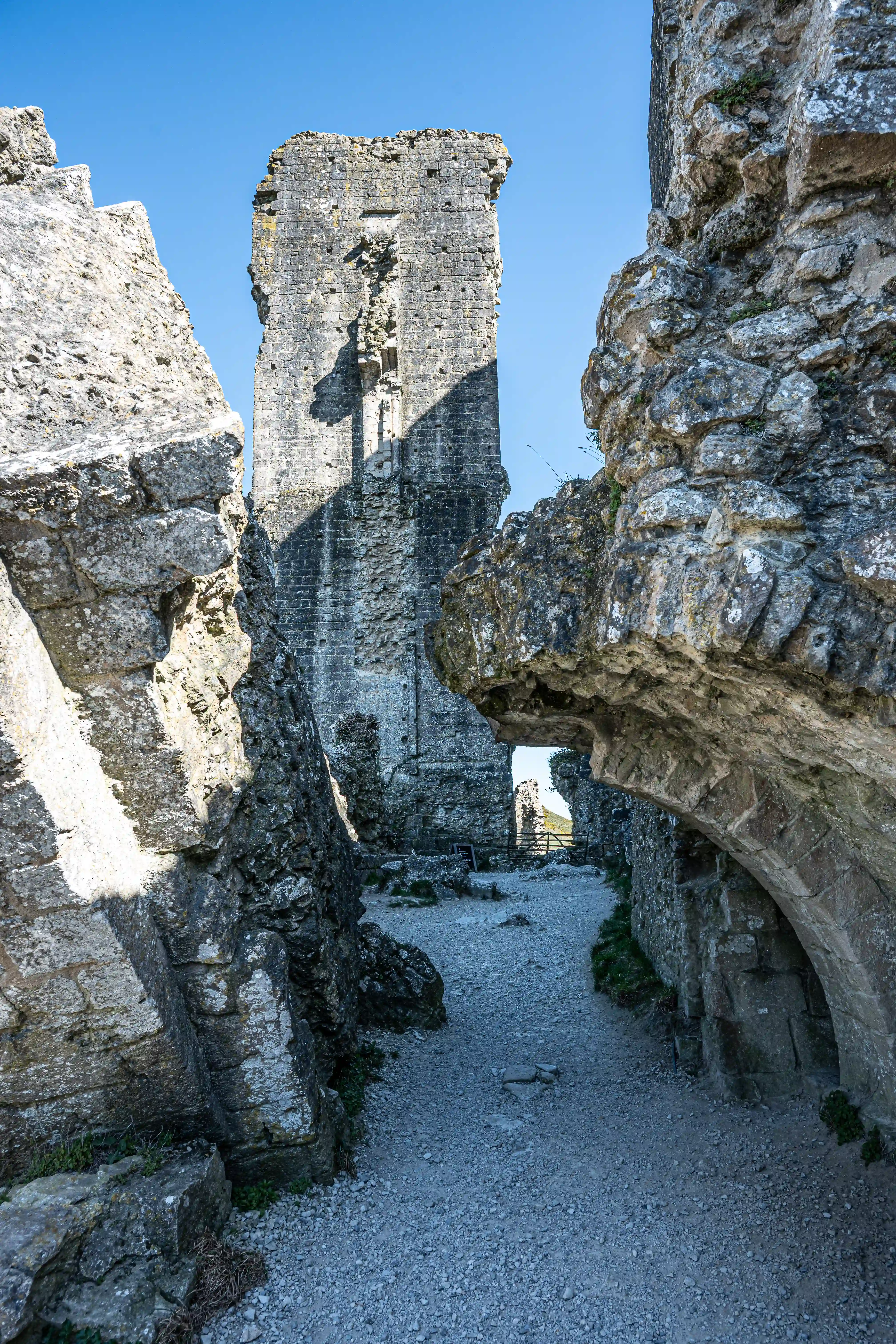 The image showcases the imposing ruins of a stone fortress, dominated by a tall, weathered tower. The structure is partially collapsed, with large sections of stone walls and tumbled rocks framing a gravel pathway. Sunlight casts strong shadows, highlighting the textures and age of the ancient architecture against a clear blue sky. The perspective draws the viewer into the ruins, suggesting a sense of exploration and history.