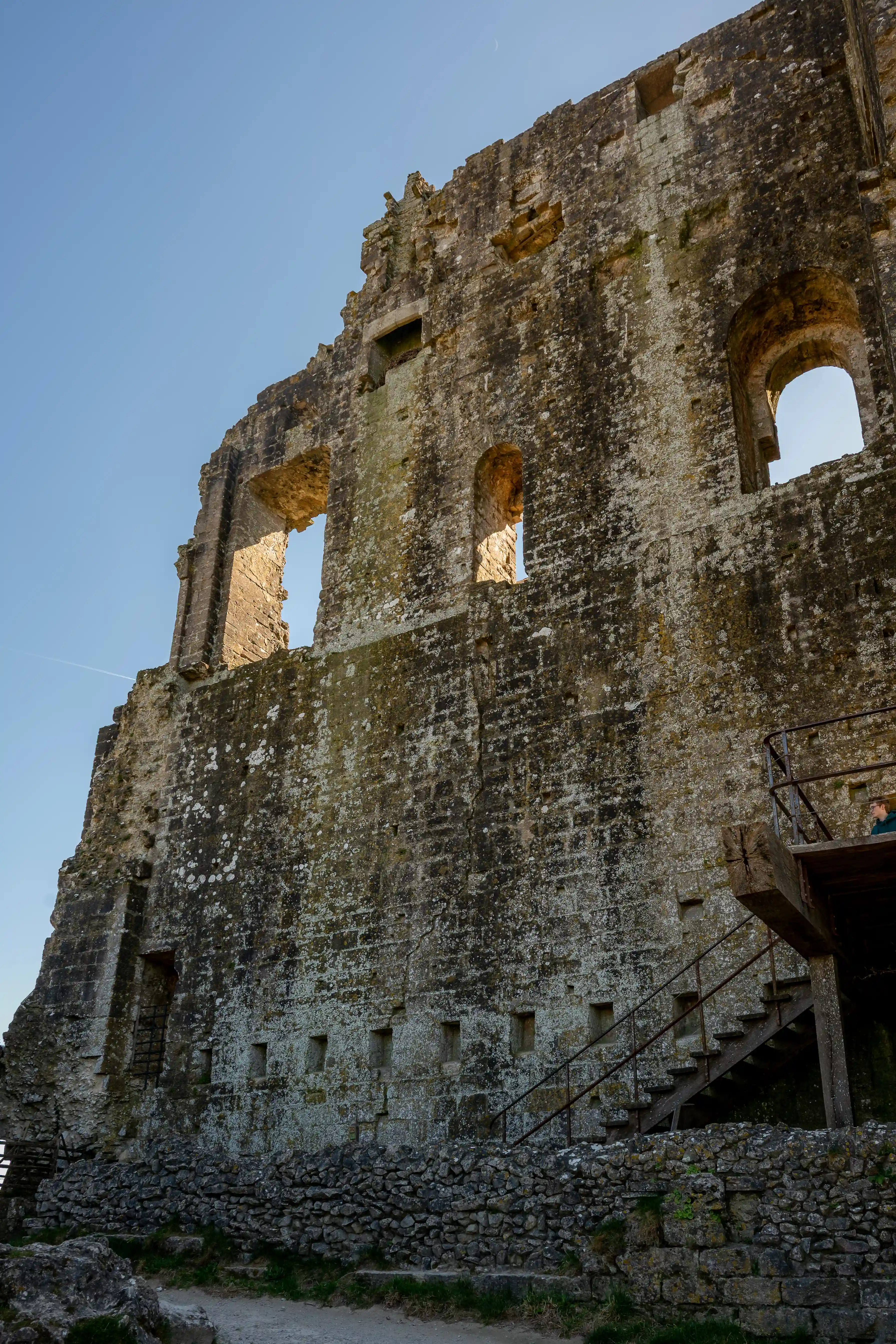 The image depicts the ruins of an ancient stone structure, likely a castle or fortress. The building shows significant wear and tear with large sections of walls missing, revealing the interior. There are several arched windows and doorways, some of which are partially collapsed. A metal staircase has been added for modern access, indicating that the site might be a historical attraction. The sky is clear, suggesting it is a sunny day.