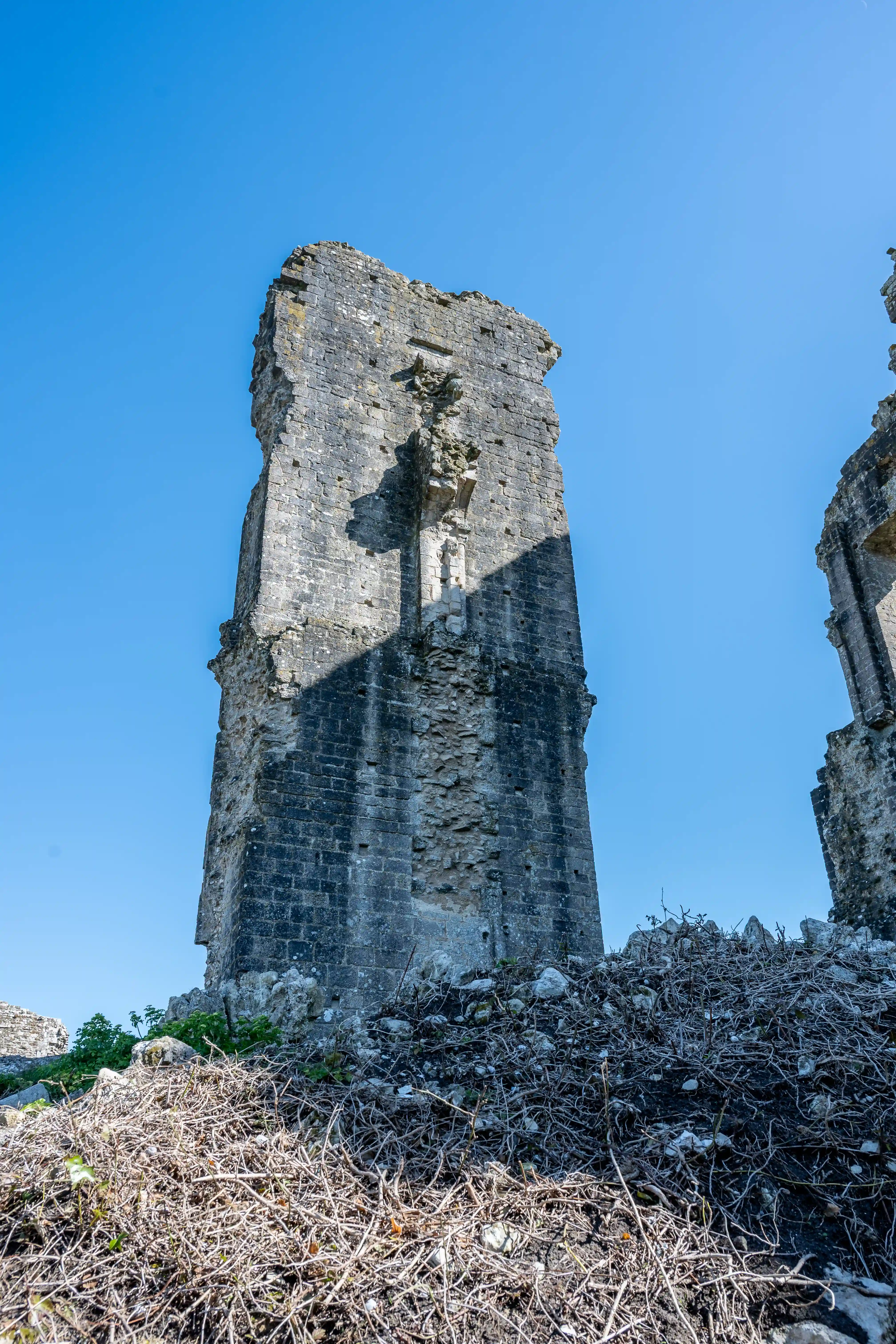 The image depicts the ruins of an old stone structure, likely a castle or fortress, with a tall, weathered tower standing prominently against a clear blue sky. The tower shows signs of significant wear and decay, with visible cracks and missing sections. The base of the tower is surrounded by dry brush and small rocks, indicating a possibly neglected or abandoned state. The overall scene suggests a historical site that has withstood the test of time, standing as a testament to its past grandeur and the passage of years.