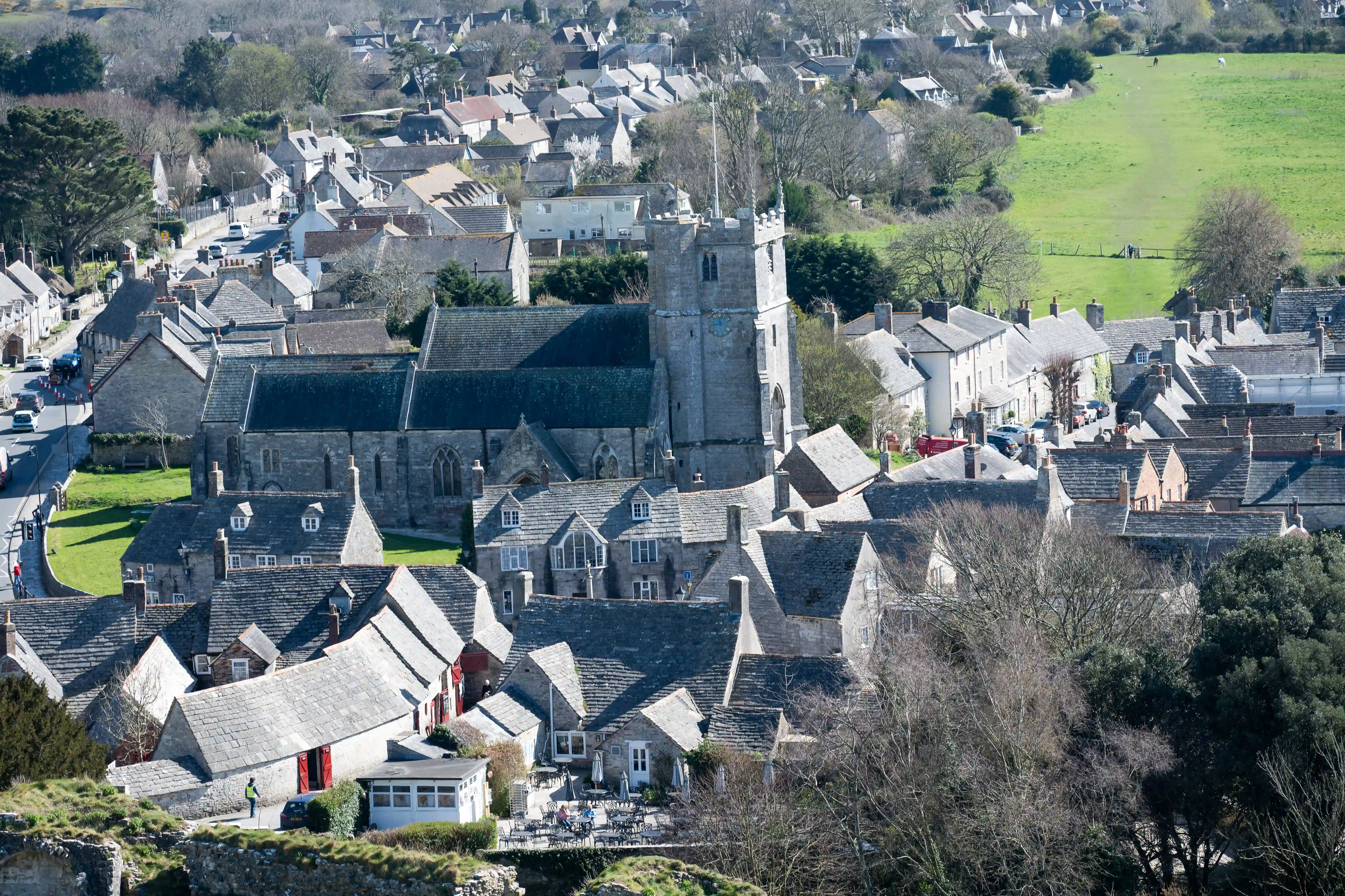 The image depicts a quaint, picturesque village with a prominent church at its center. The church features a tall tower with a clock and is surrounded by closely packed stone houses with slate roofs. The village is nestled in a lush, green landscape with open fields and scattered trees. The streets are narrow and winding, with a few cars and pedestrians visible. The overall atmosphere is serene and charming, typical of a traditional countryside village.