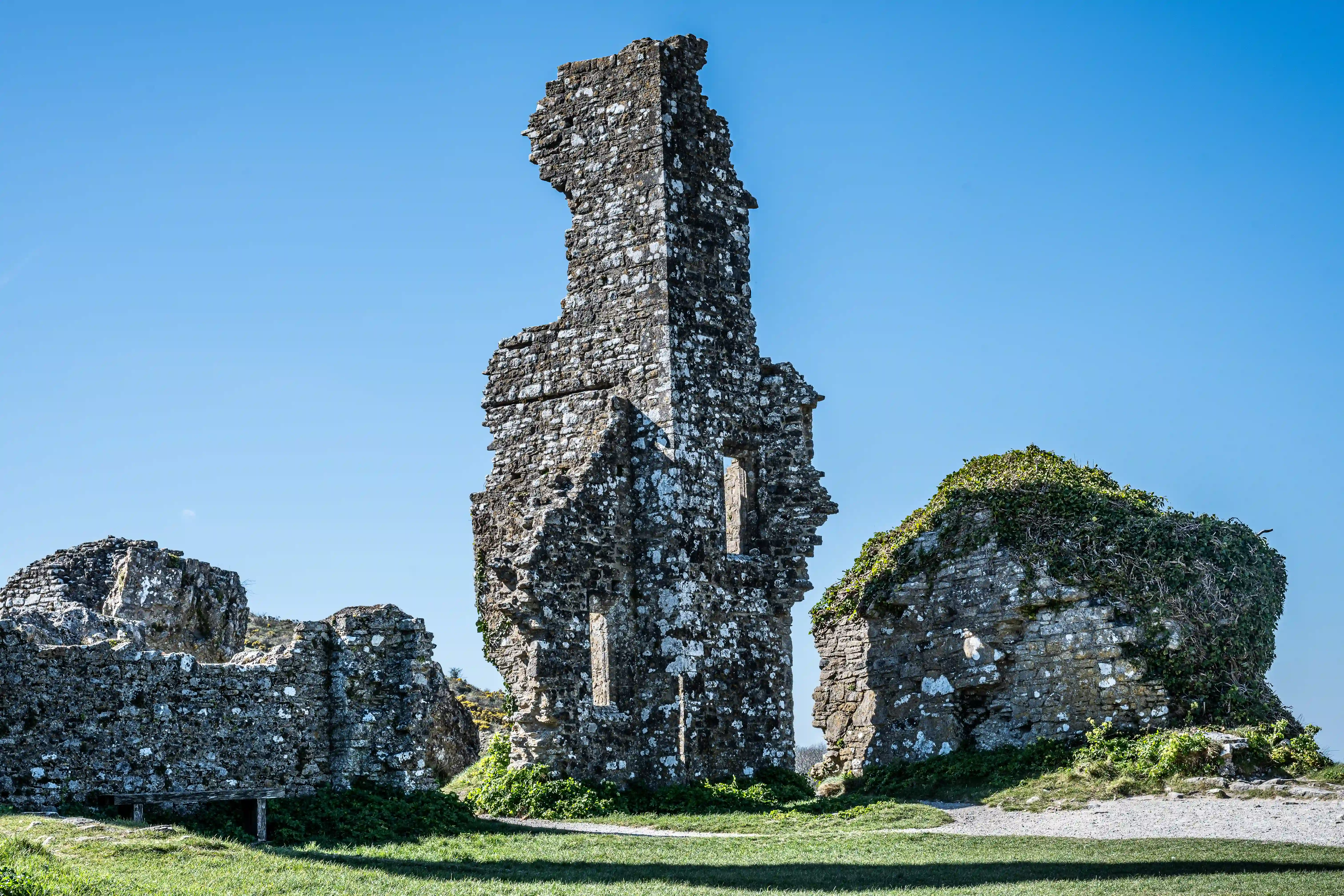 The image depicts the ruins of an old stone structure, likely a castle or fortification, with a central tower and surrounding walls. The structure is heavily weathered and covered in patches of greenery, indicating it has been abandoned for a significant period. The sky above is clear and blue, suggesting a sunny day. The ground around the ruins is grassy, with a dirt path leading up to the entrance of the structure. The overall scene conveys a sense of historical significance and natural reclamation.