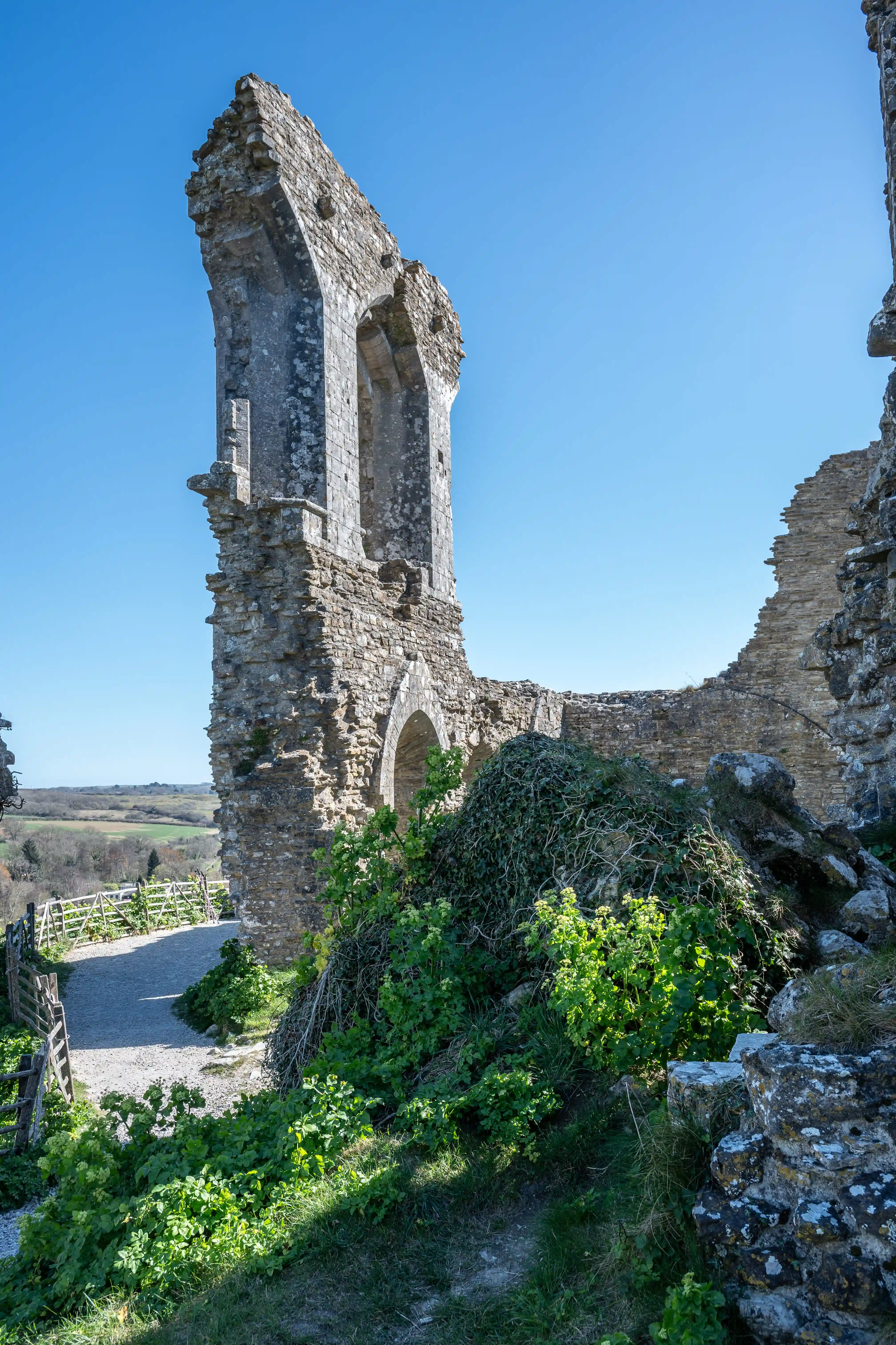 The image depicts the ruins of an old stone structure, possibly a castle or fortification, with partially standing walls and arches. The ruins are surrounded by greenery and situated on elevated ground, overlooking a landscape of fields and distant hills under a clear blue sky.