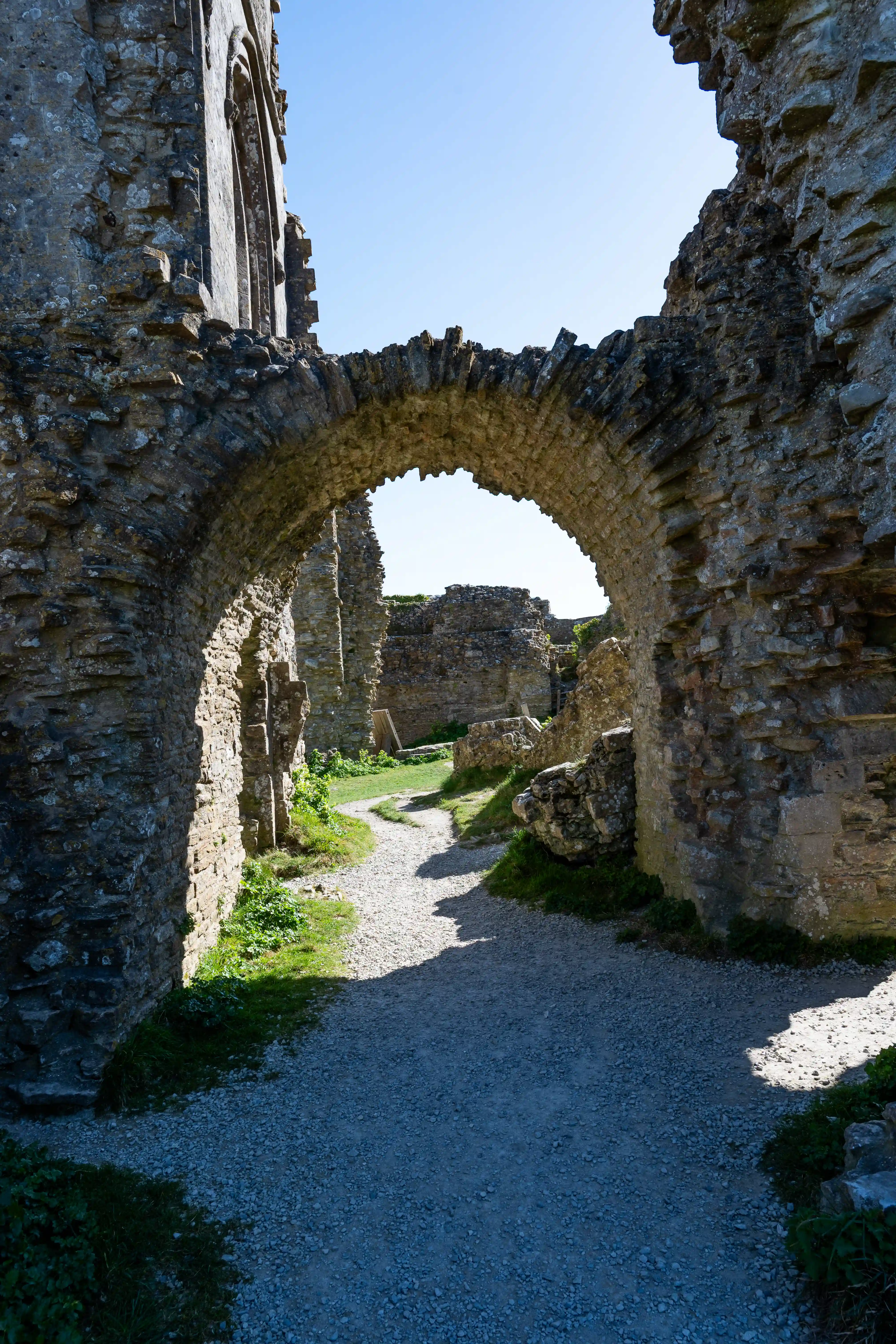 The image depicts the ruins of an old stone structure, likely a castle or fortress. The pathway through the ruins is framed by two large, weathered stone arches. The ground is covered with gravel and patches of grass, leading further into the ruins. The sky is visible through the arches, indicating an open area beyond the immediate structure. The overall scene conveys a sense of historical significance and architectural grandeur.