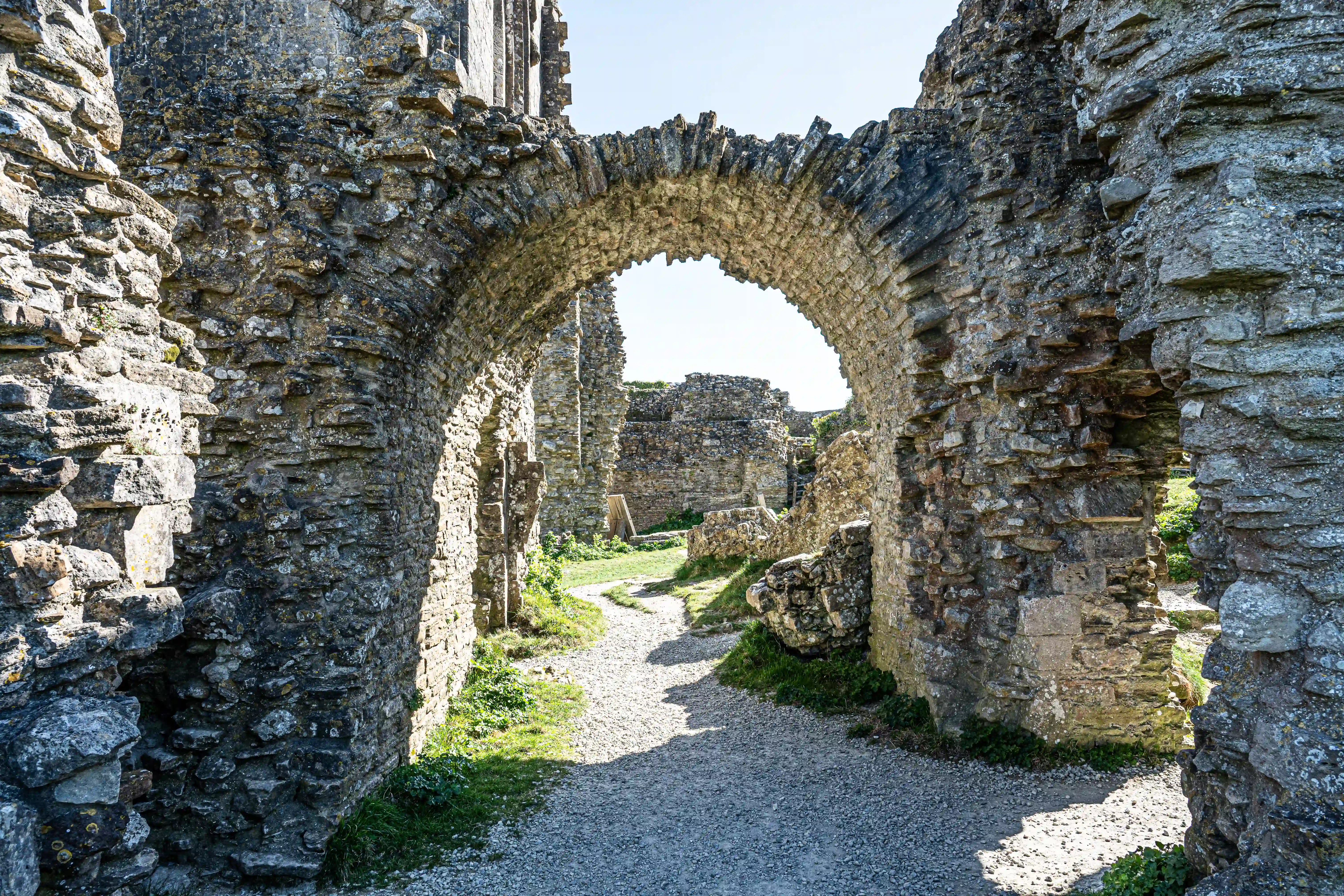 The image depicts the ruins of an old stone structure, likely a castle or fortification. The pathway through the ruins is bordered by weathered stone walls and arches, leading further into the remains of the building. The scene is bathed in natural light, highlighting the texture and age of the stones. Vegetation is growing in patches along the ground, indicating a degree of neglect and the passage of time. The overall atmosphere is one of historical significance and quiet decay.