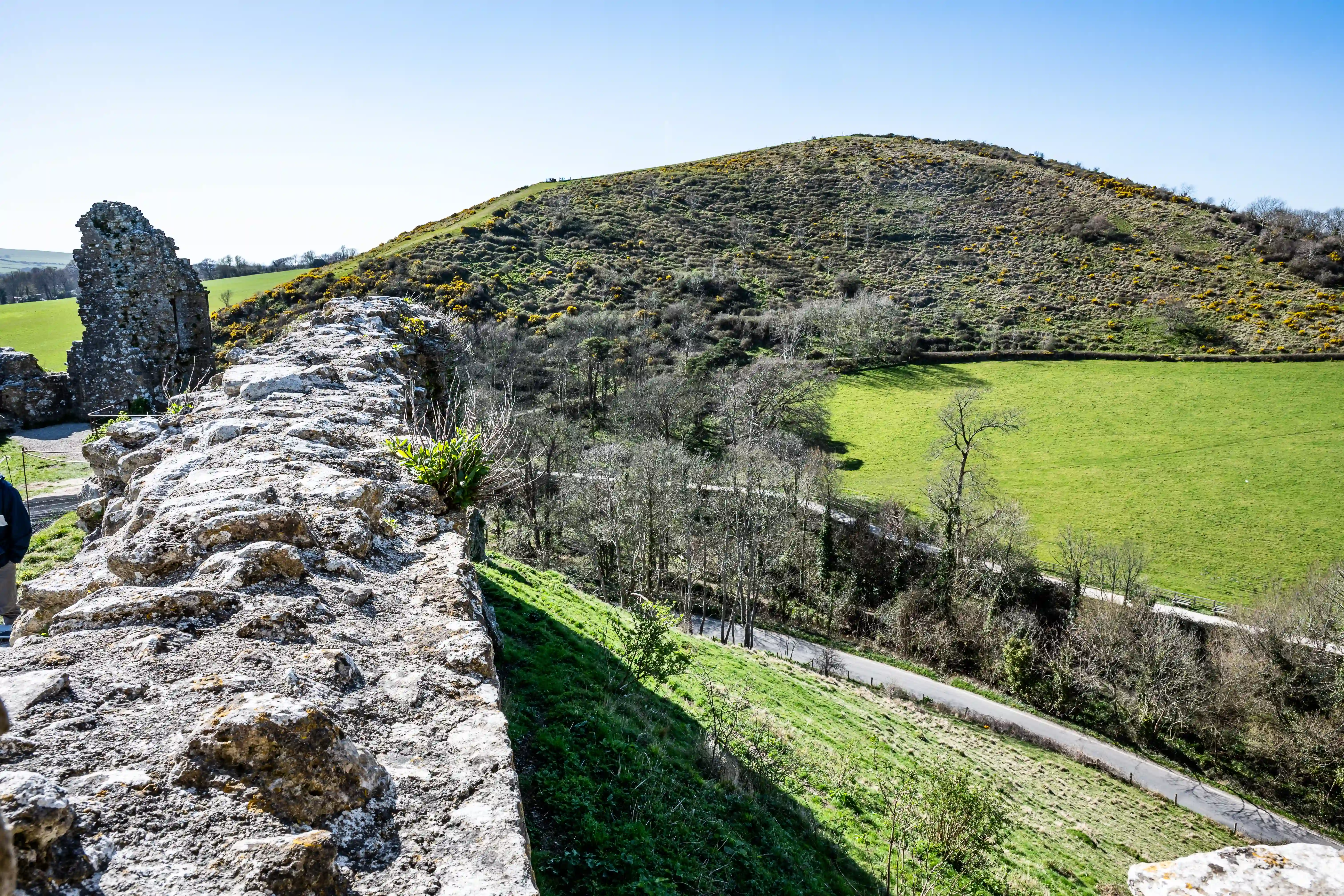 The image depicts the ruins of an ancient structure on a hillside, surrounded by lush greenery and a clear blue sky. The ruins consist of weathered stone walls and pathways, with a mix of vegetation growing around them. The scene suggests a historical site, possibly a castle or fort, situated in a rural, scenic landscape.