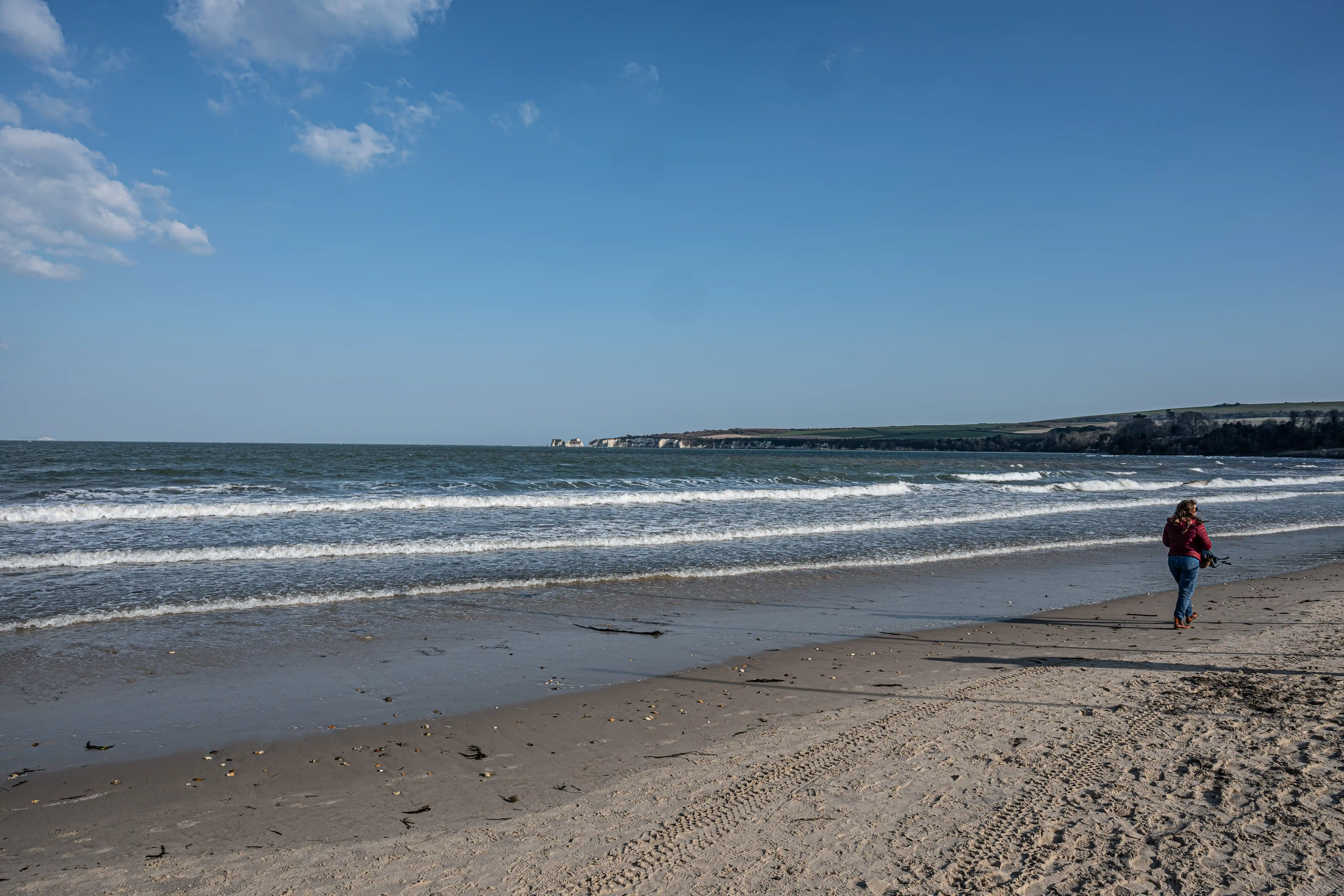 The image depicts a serene beach scene with gentle waves, a clear sky, and a person walking along the shore. The beach has a mix of sand and small pebbles, and the coastline extends into the distance with some rocky formations. The overall atmosphere is calm and peaceful, suggesting a pleasant day by the sea.