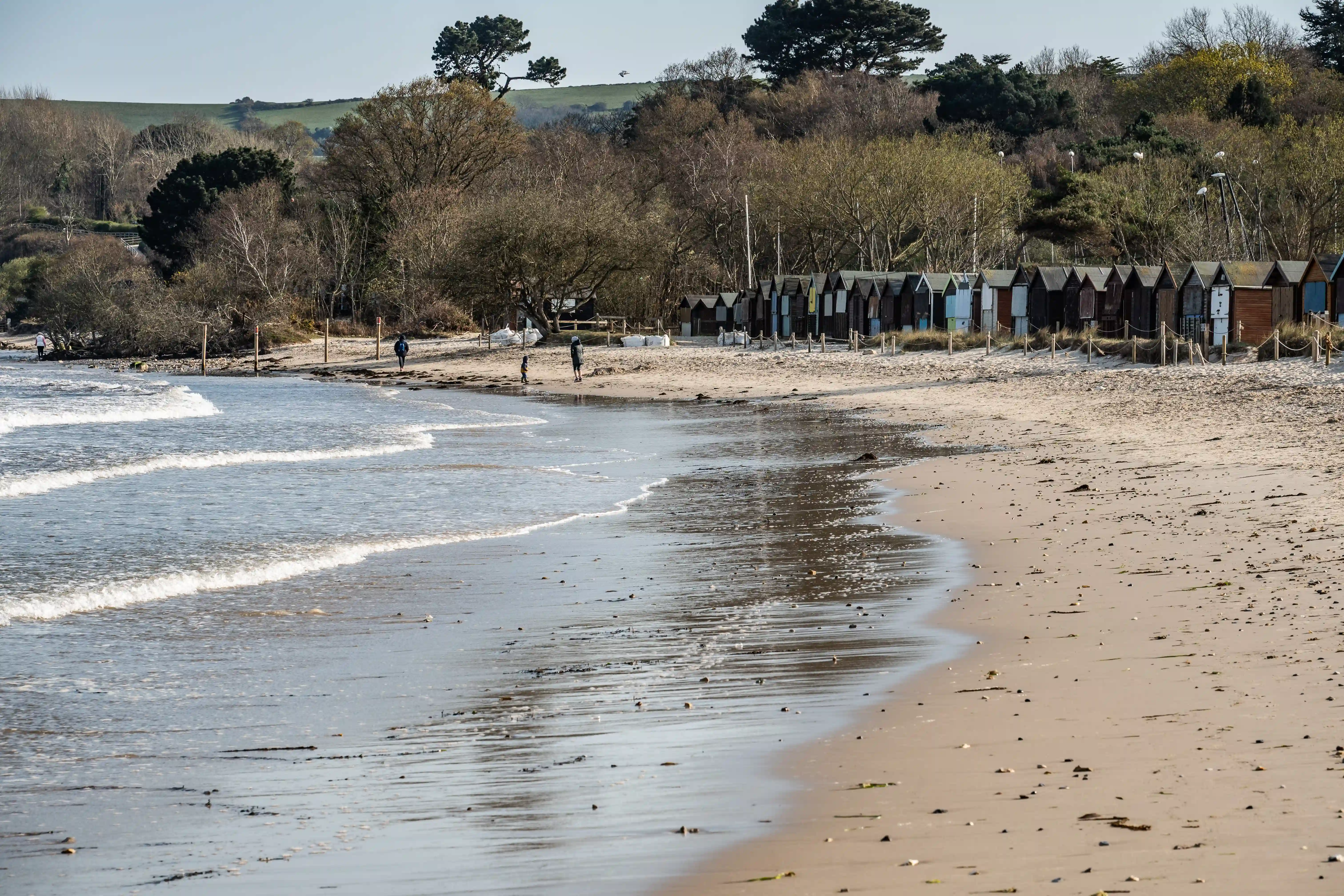 The image depicts a serene beach scene with a few people walking along the shoreline. The beach is bordered by a row of colorful beach huts and a backdrop of trees and rolling hills. The tide is out, revealing wet sand and shallow pools of water. The overall atmosphere is calm and peaceful, suggesting a quiet day at the beach.