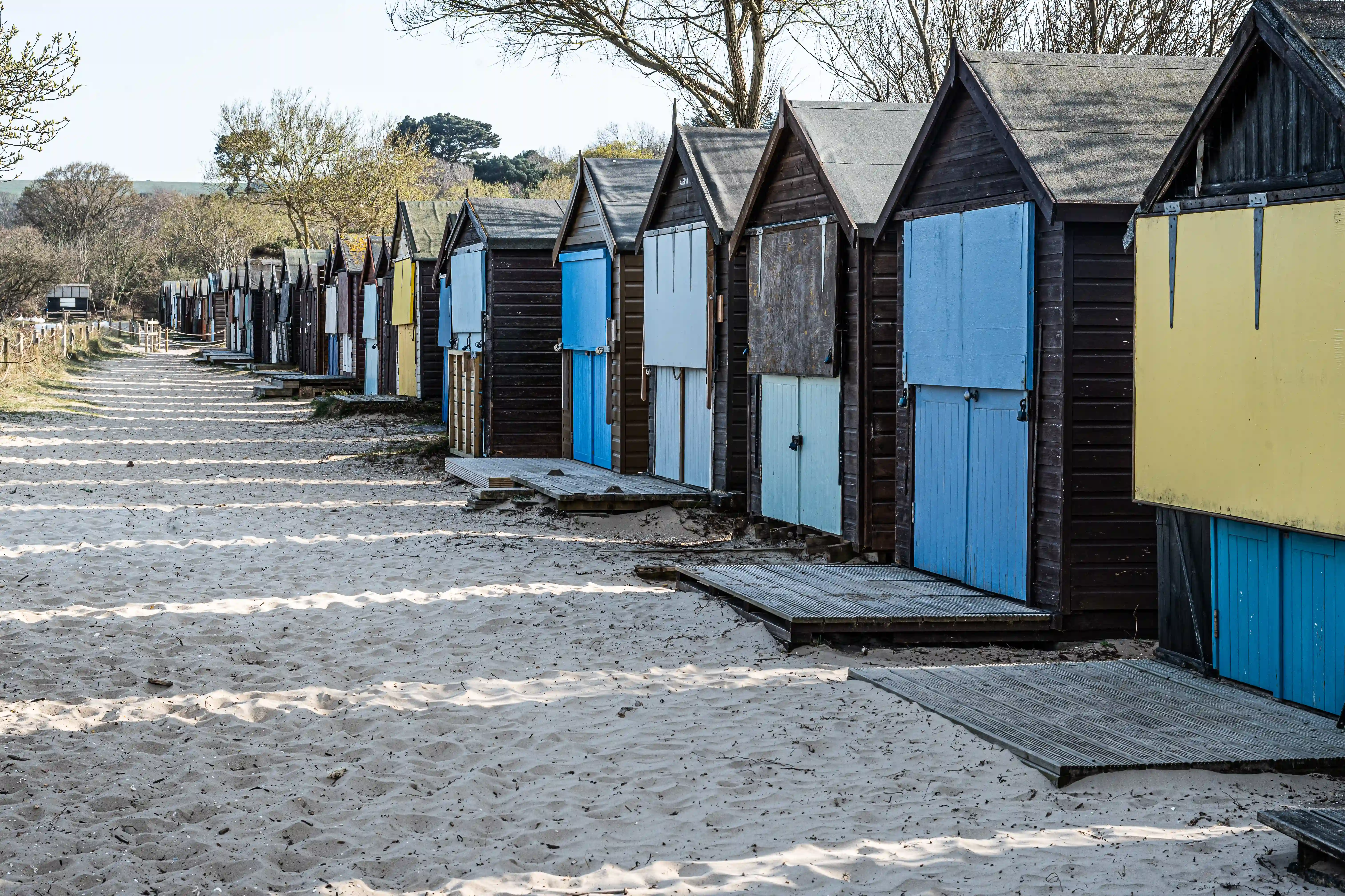 The image shows a row of beach huts lined up on a sandy beach. The huts are made of wood and have various colored doors, including blue, yellow, and white. Each hut has a small porch with steps leading up to it. The beach appears to be quiet and serene, with no people visible in the image. In the background, there are trees and a hilly landscape.